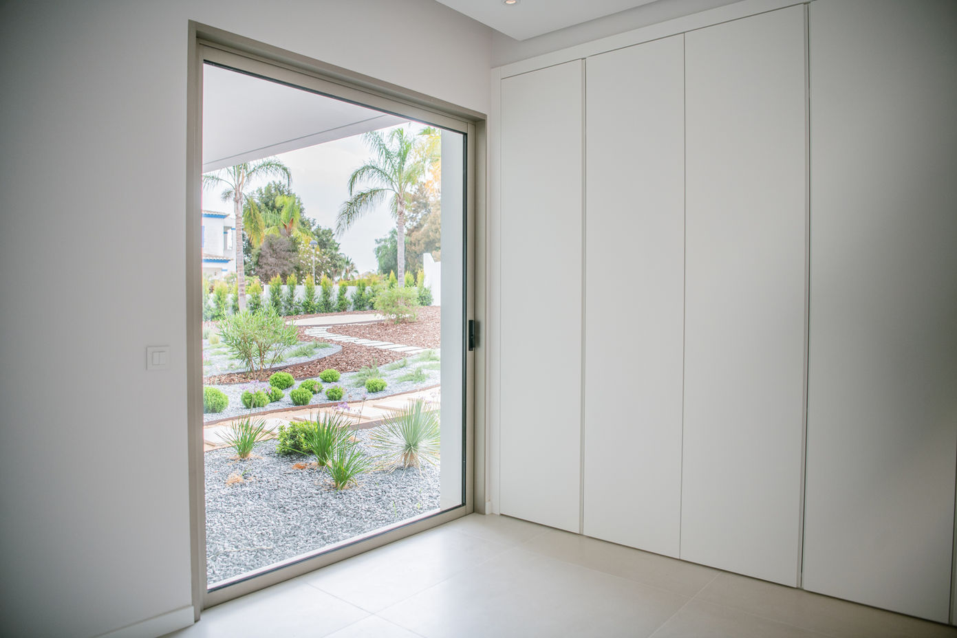29bf52_af4a61e7a2f646f595df513e18160704-mv2 Modern interior with white built-in wardrobes and a large sliding glass door opening to a desert garden with palm trees.