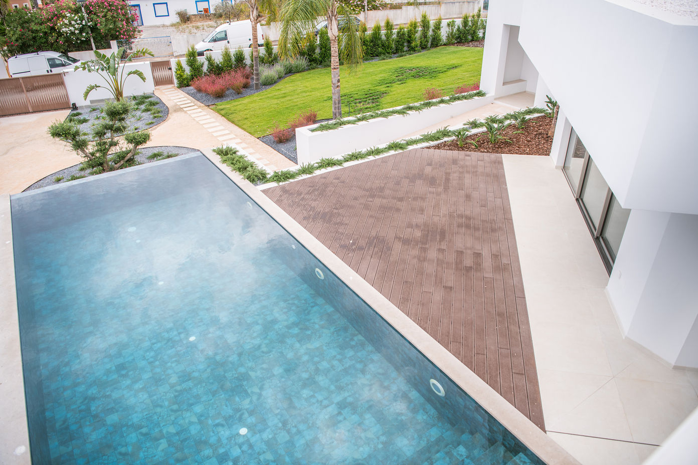 29bf52_b0442c897b4f48bb9b31e816d622714f-mv2 Contemporary backyard with a rectangular blue pool, brown wooden deck, and a white modern house façade.