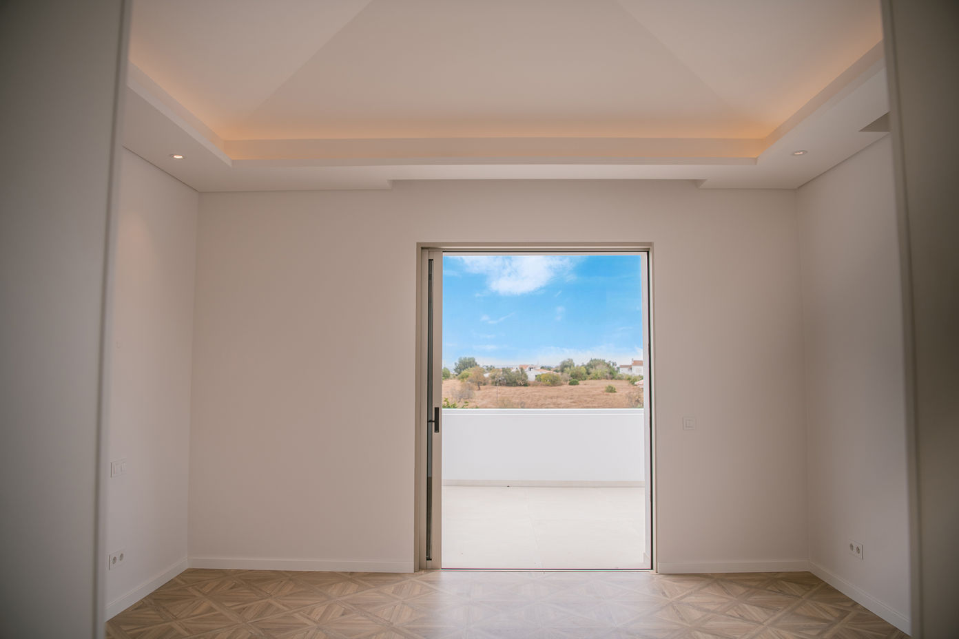 29bf52_bcfb5949b3ad4abcba5d8e3950187041-mv2 Bright, empty room with white walls and a sliding glass door opening to a balcony; blue sky and distant hills visible outside.