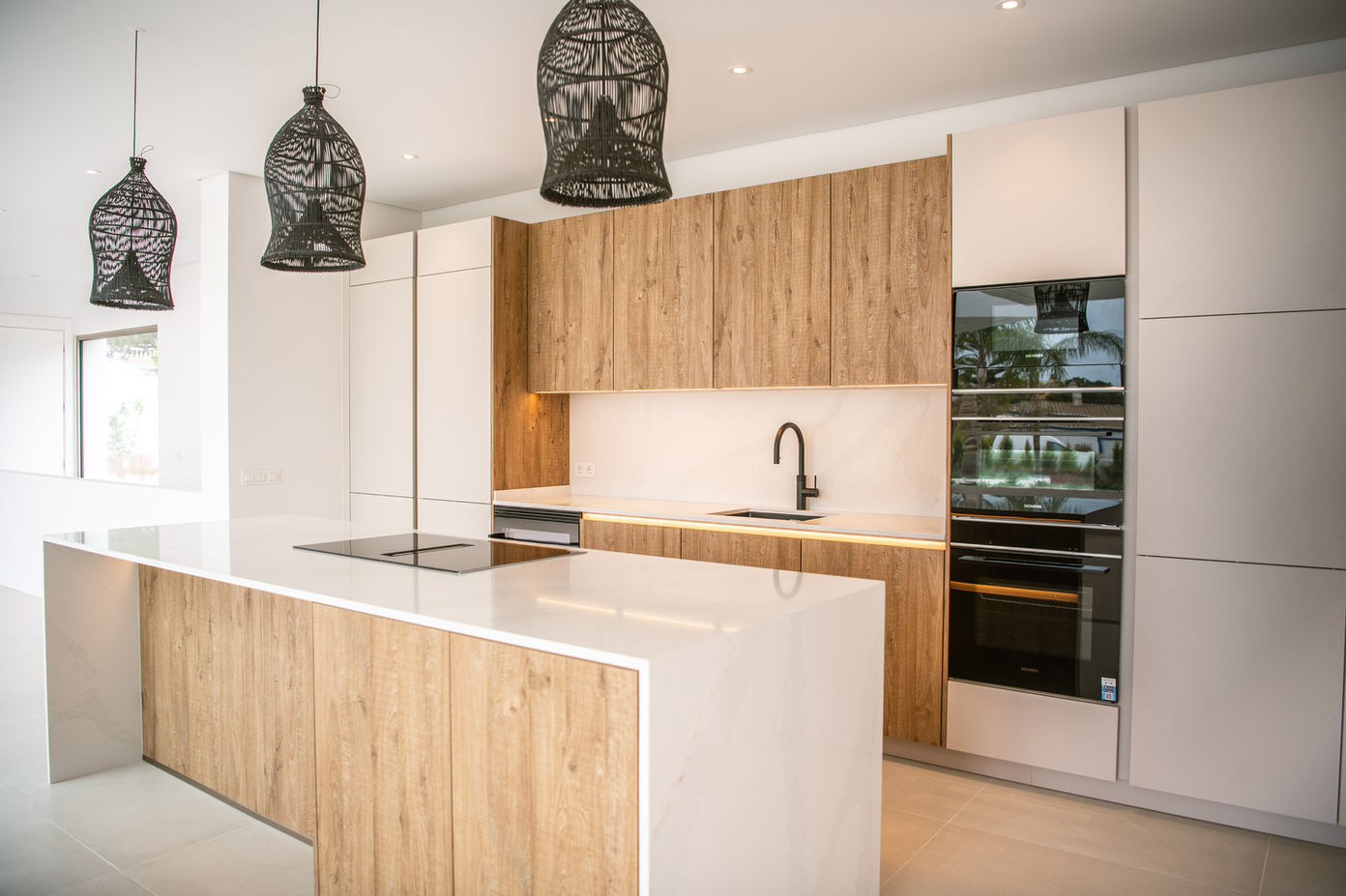 29bf52_de07ab28b63c4ae7bba1ae31343b86bb-mv2 Modern kitchen with white countertops and a large island, wood cabinets, a black faucet, and built‑in ovens on the right.