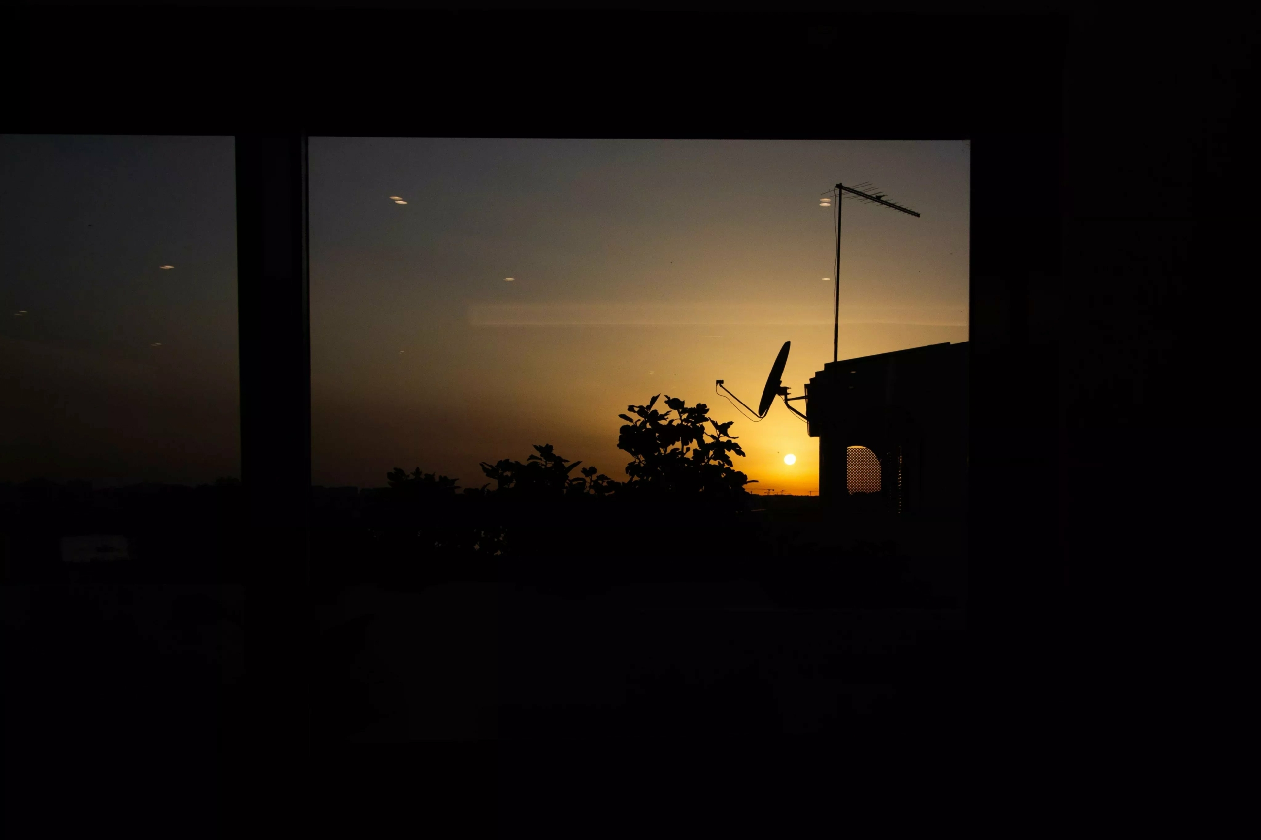 Sunset skyline seen through a window, with a rooftop antenna silhouetted against the orange sky on the horizon.