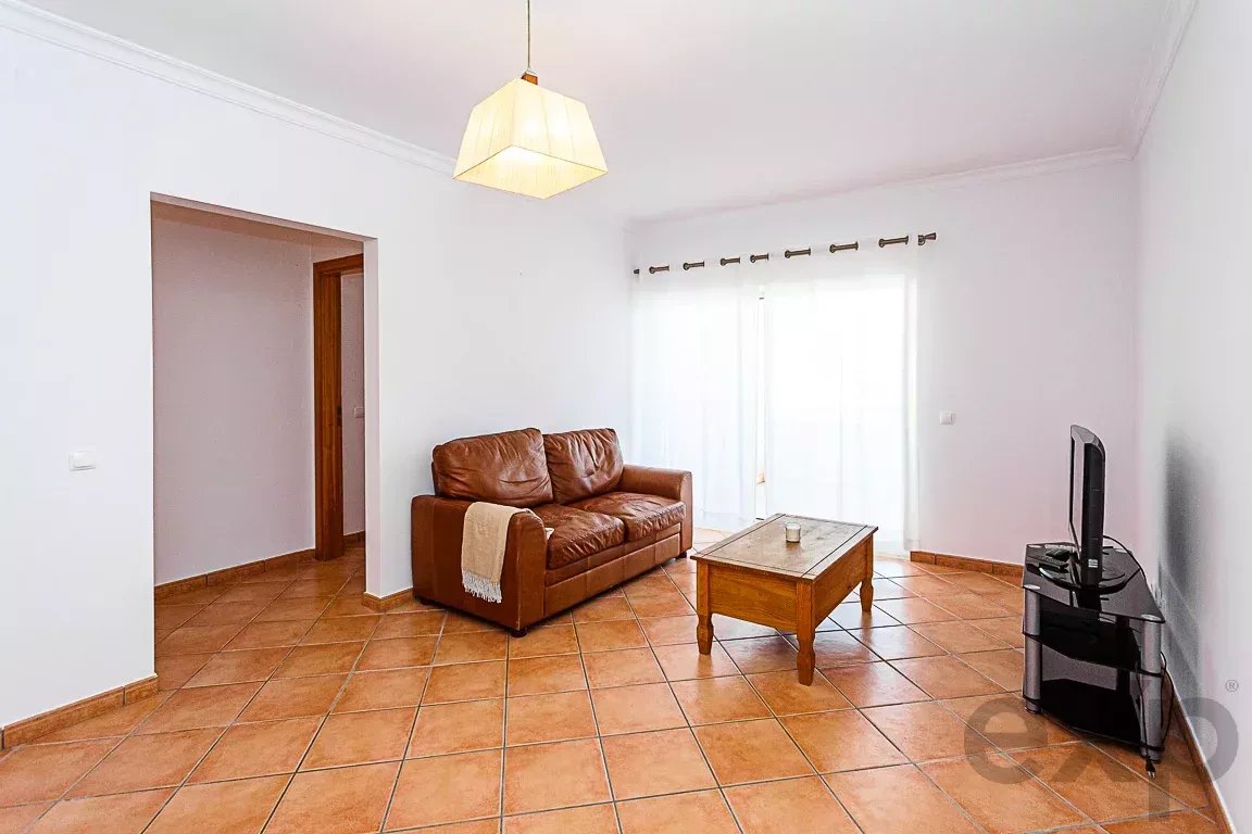 Cozy living room with a brown leather sofa, throw blanket, wooden coffee table, and a TV on a glass stand near white paneled walls and tiled floor.