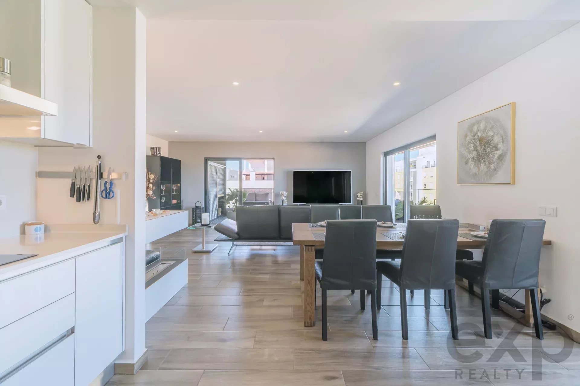 Open-plan living and dining area with white kitchen on the left, gray dining chairs, and a large TV opposite.|Modern bright home interior featuring a kitchen, dining table for eight, and a gray sofa facing a wall-mounted TV.|Contemporary apartment open space with a dining table, gray seating, large windows, and wall art.|Bright, airy living-dining area with white cabinetry, wooden table, and a mounted flat-screen TV across from seating.