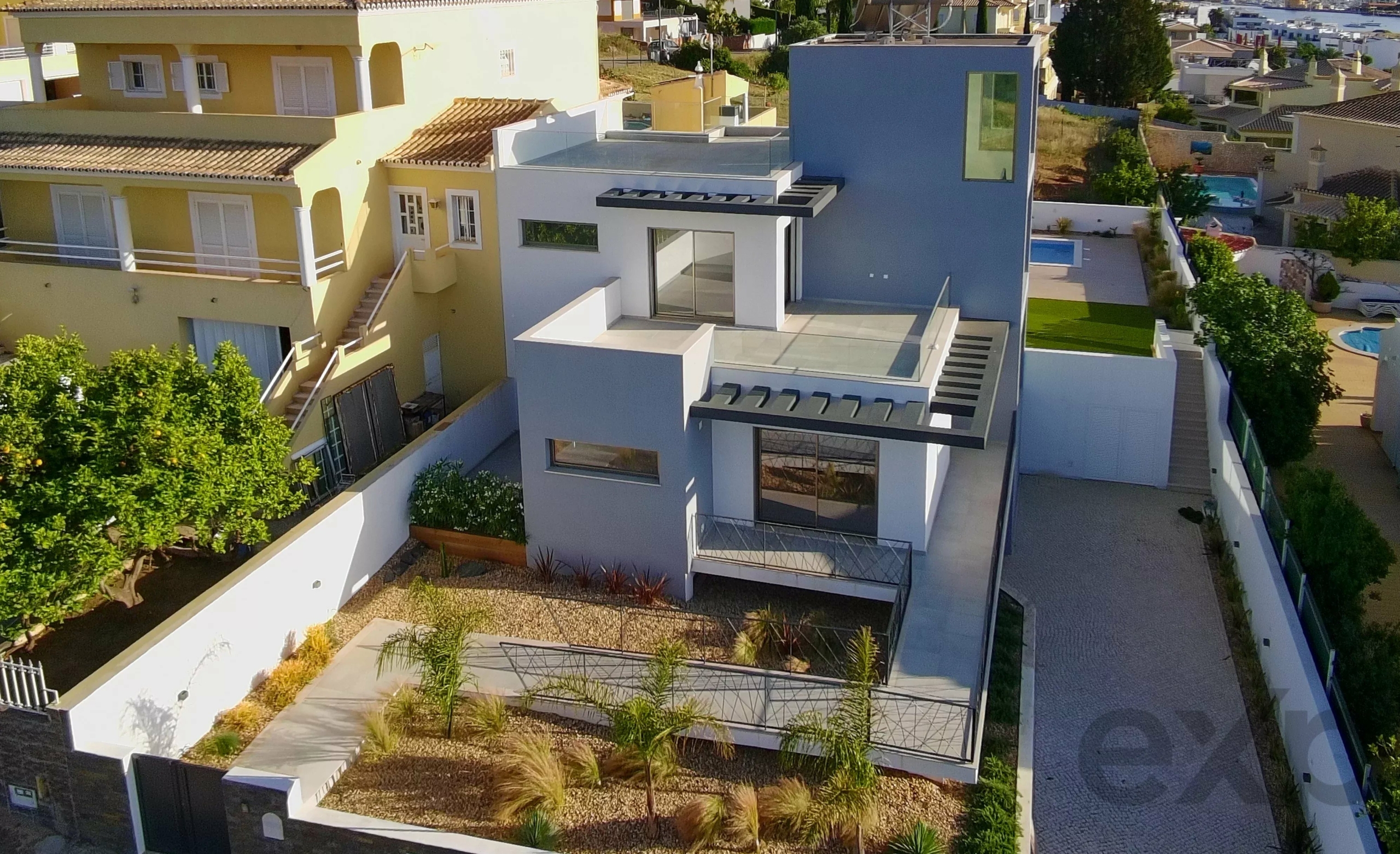 Aerial view of a modern two-tone house (white and blue) with glass railings, balconies, and a paved driveway nearby in a sunny neighborhood.