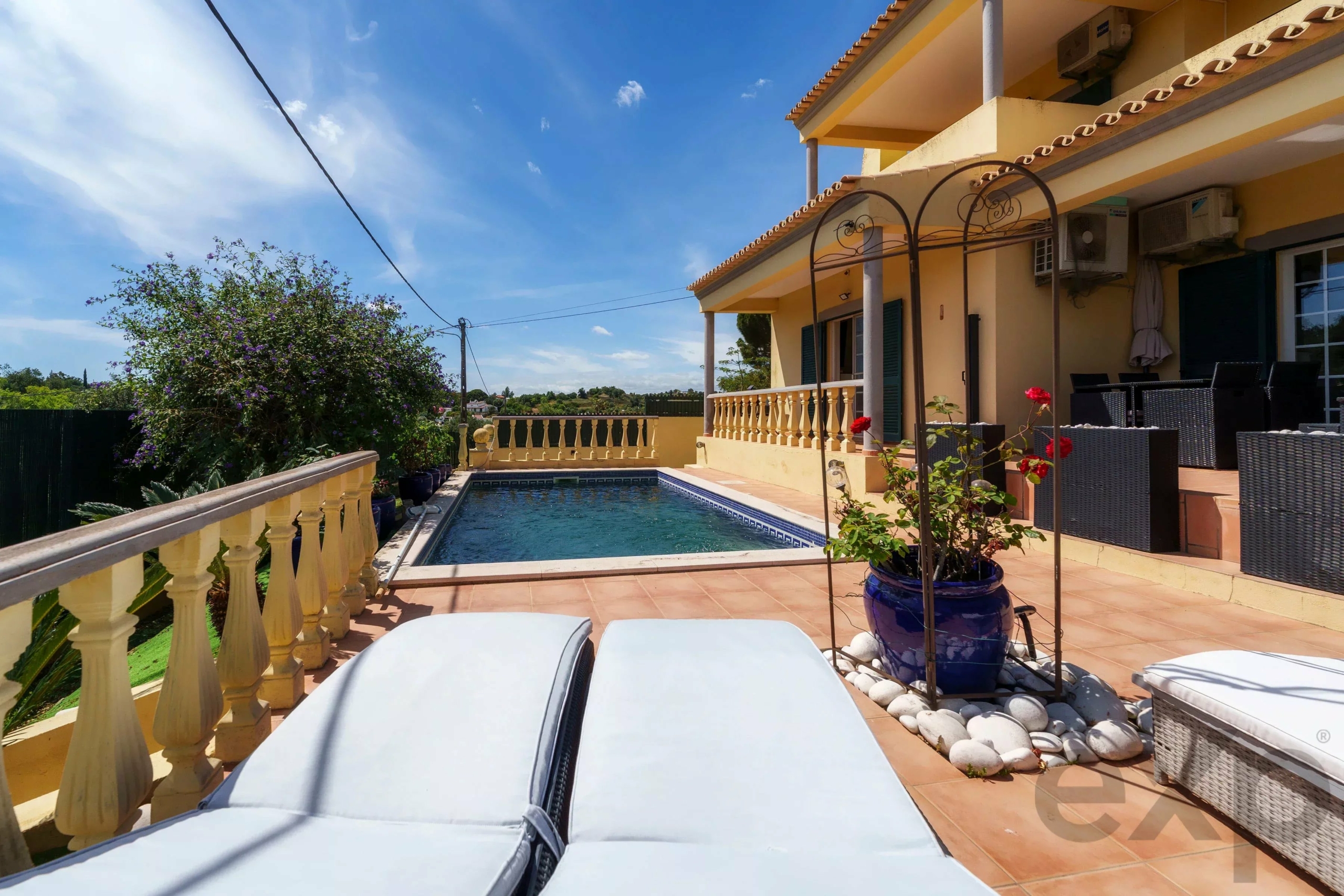 Sunny backyard patio with a rectangular pool, beige railings, and cream-colored loungers facing the water.