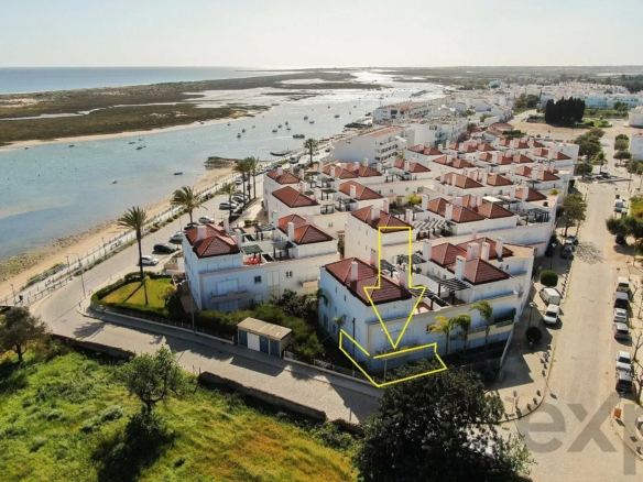Aerial view of a coastal town with white buildings and red-tiled roofs along a waterway and beach, with boats in the canal.