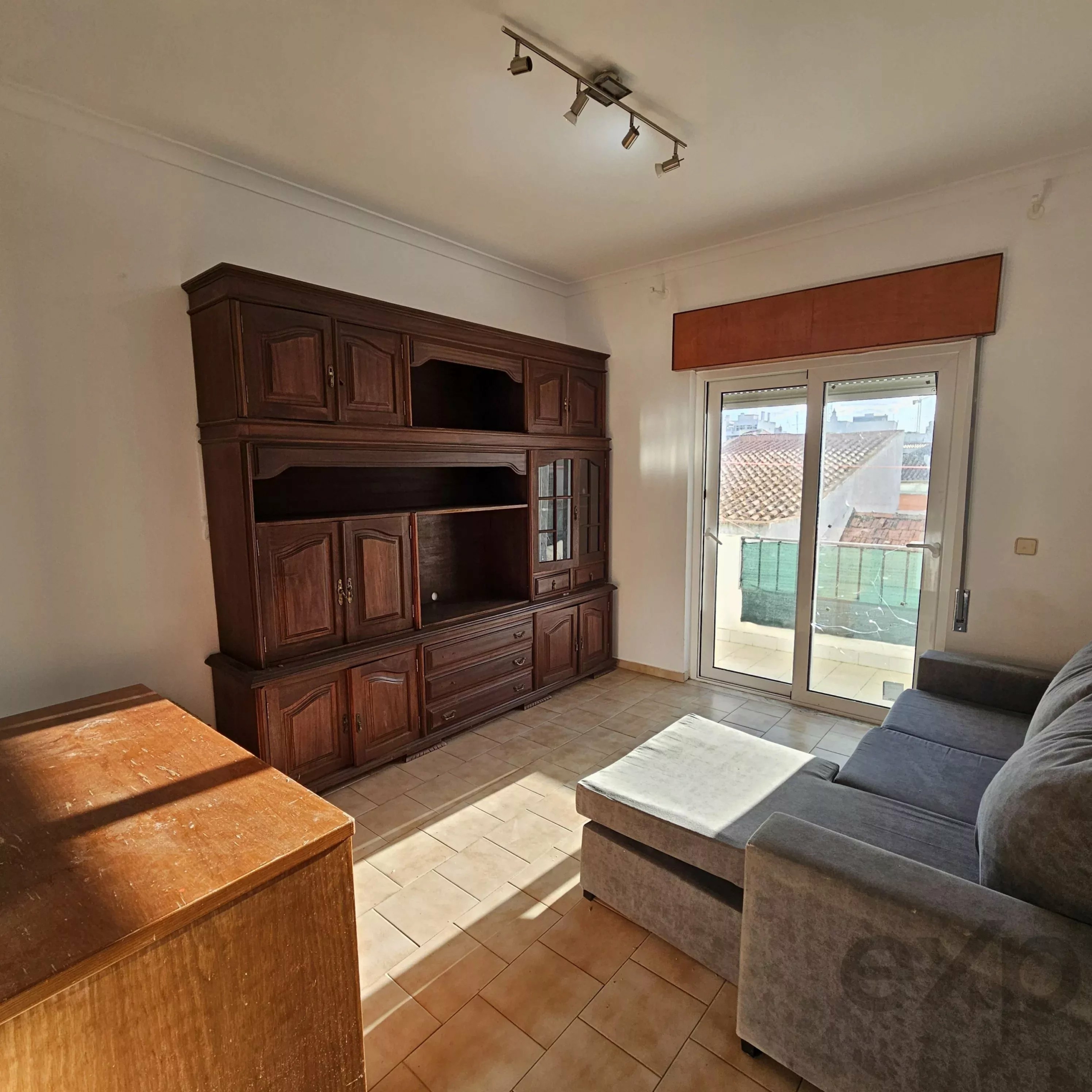 Corner of a living room with a tall dark wood cabinet, tiled floor, and a gray sectional sofa by a glass door to a balcony/or outside.