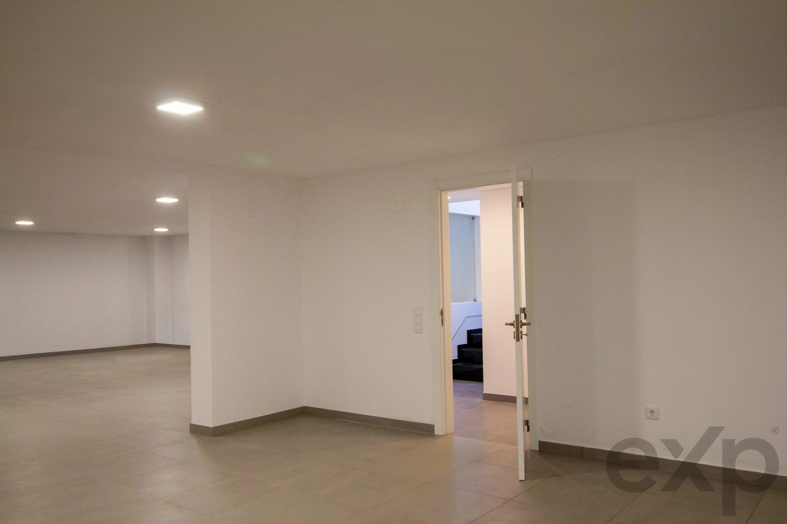 Empty, spacious room with white walls and tiled floor; an open door leads to another area.