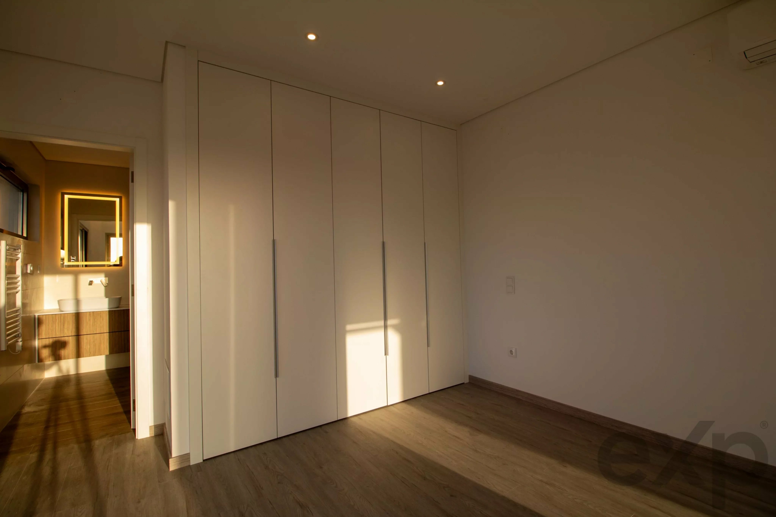 Line of tall white wardrobes along a wall in a bright, modern bedroom with warm light from a bathroom mirror visible on the left.