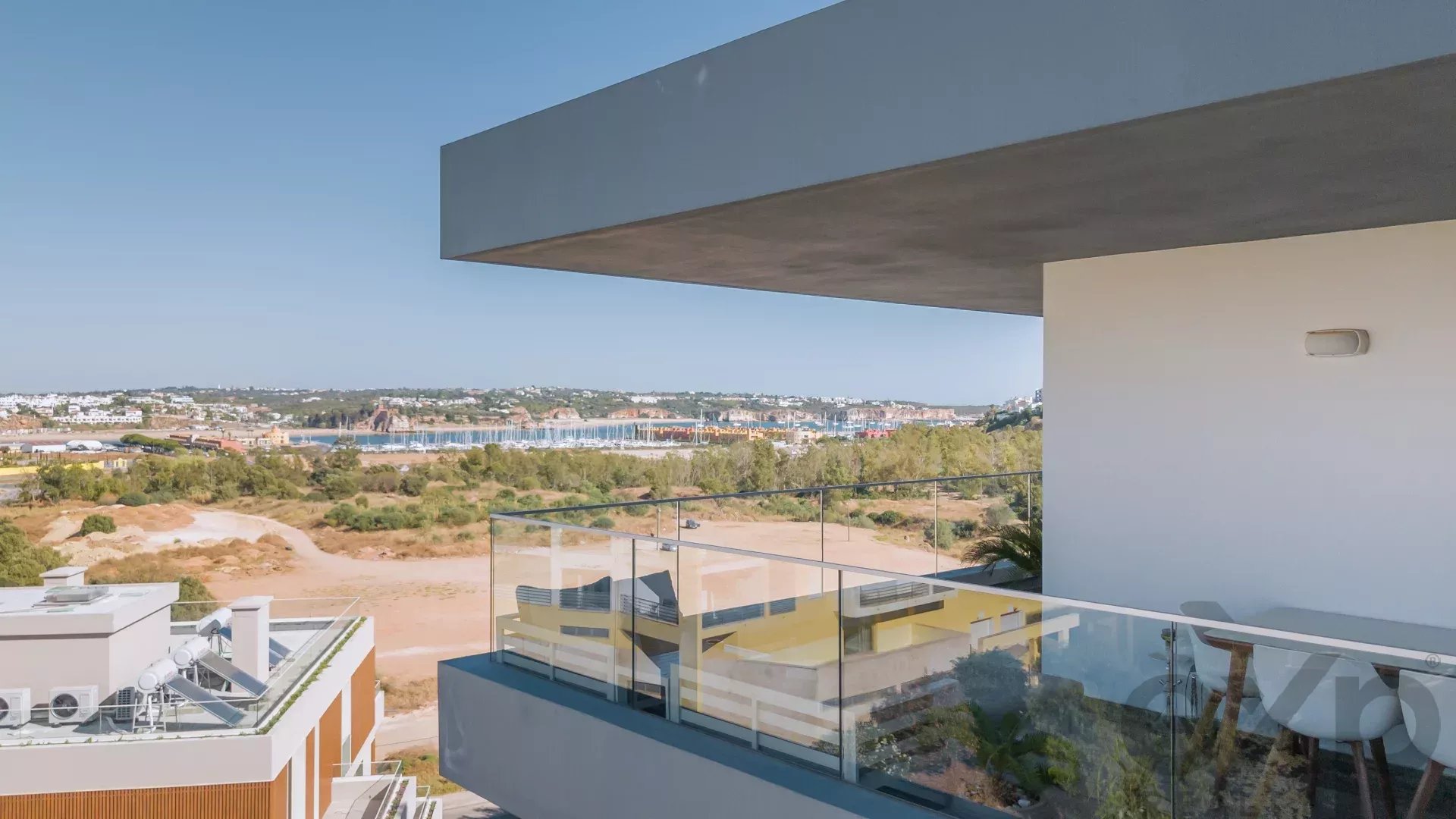 Cantilevered balcony with glass railing, overlooking a coastal town and marina under a clear blue sky.
