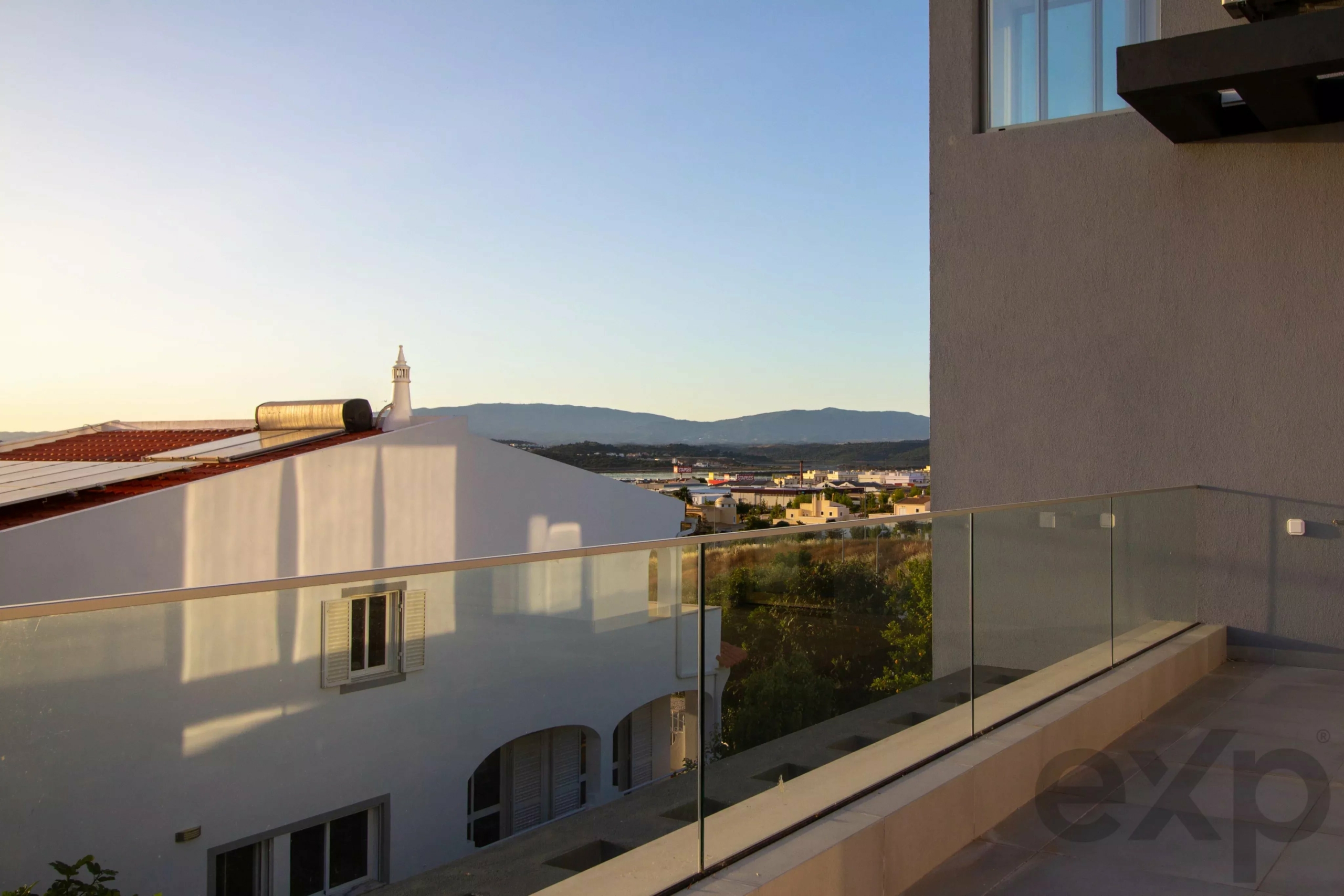 Modern balcony with glass railing overlooking a white building, red-tiled roof, and distant mountains under a clear sky at sunset/sunrise
