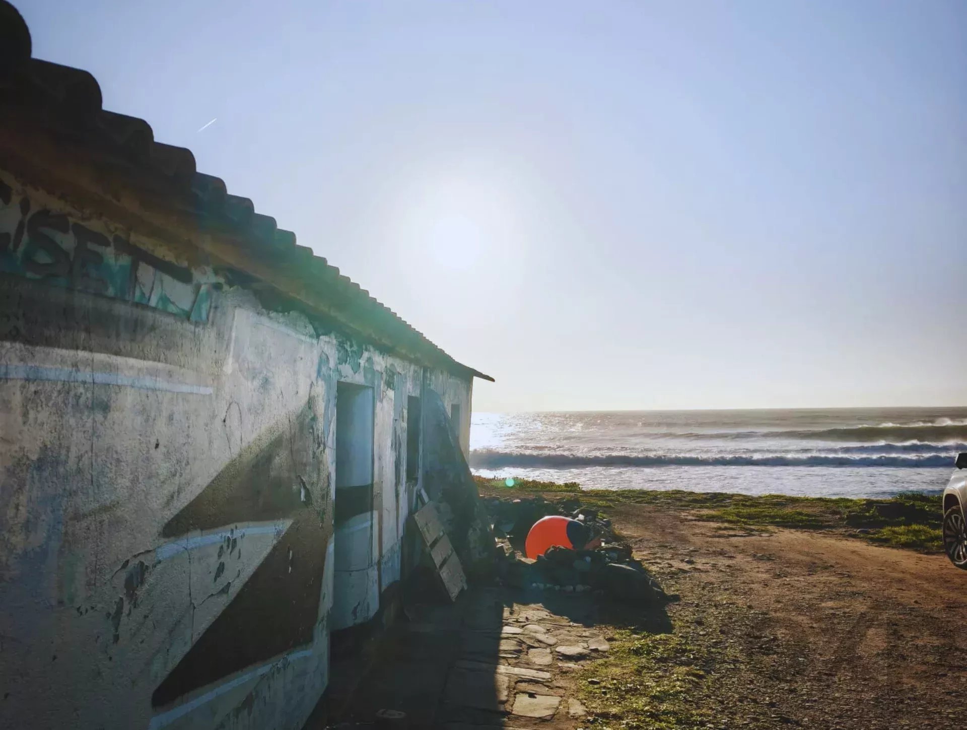 Weathered seaside shack with a tiled roof along a rocky beach, sun over a calm sea.