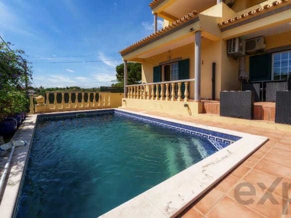 Poolside view of a yellow house with a tiled balcony, orange patio, and blue shutters under a clear sky.