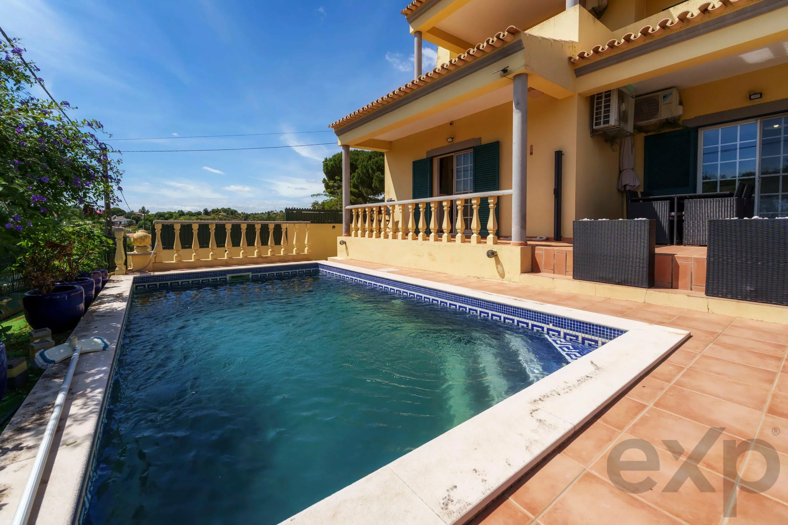 Poolside view of a yellow house with a tiled balcony, orange patio, and blue shutters under a clear sky.