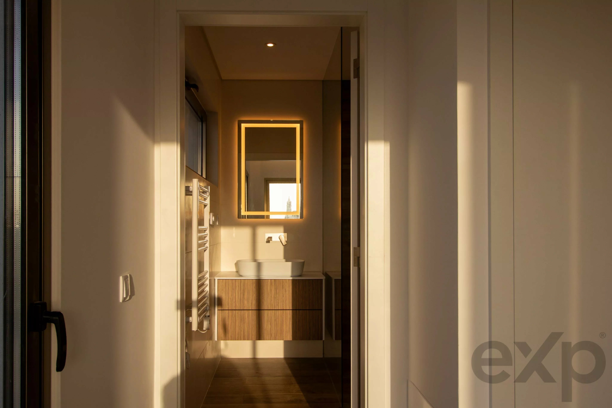 Bathroom with wooden vanity, vessel sink, and backlit square mirror; warm lighting.