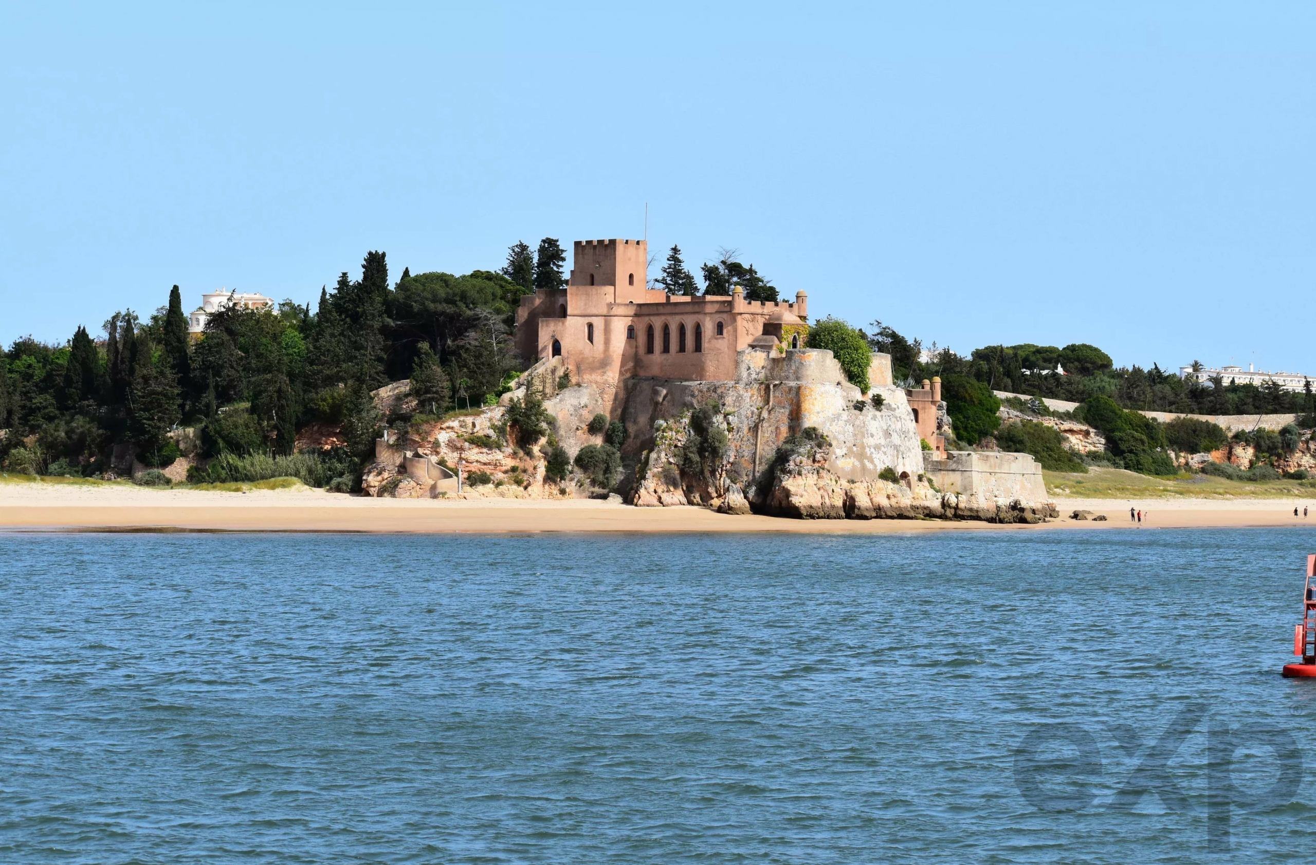 Historic fortress atop a rocky promontory overlooking a sandy beach and calm blue sea, with green trees behind.