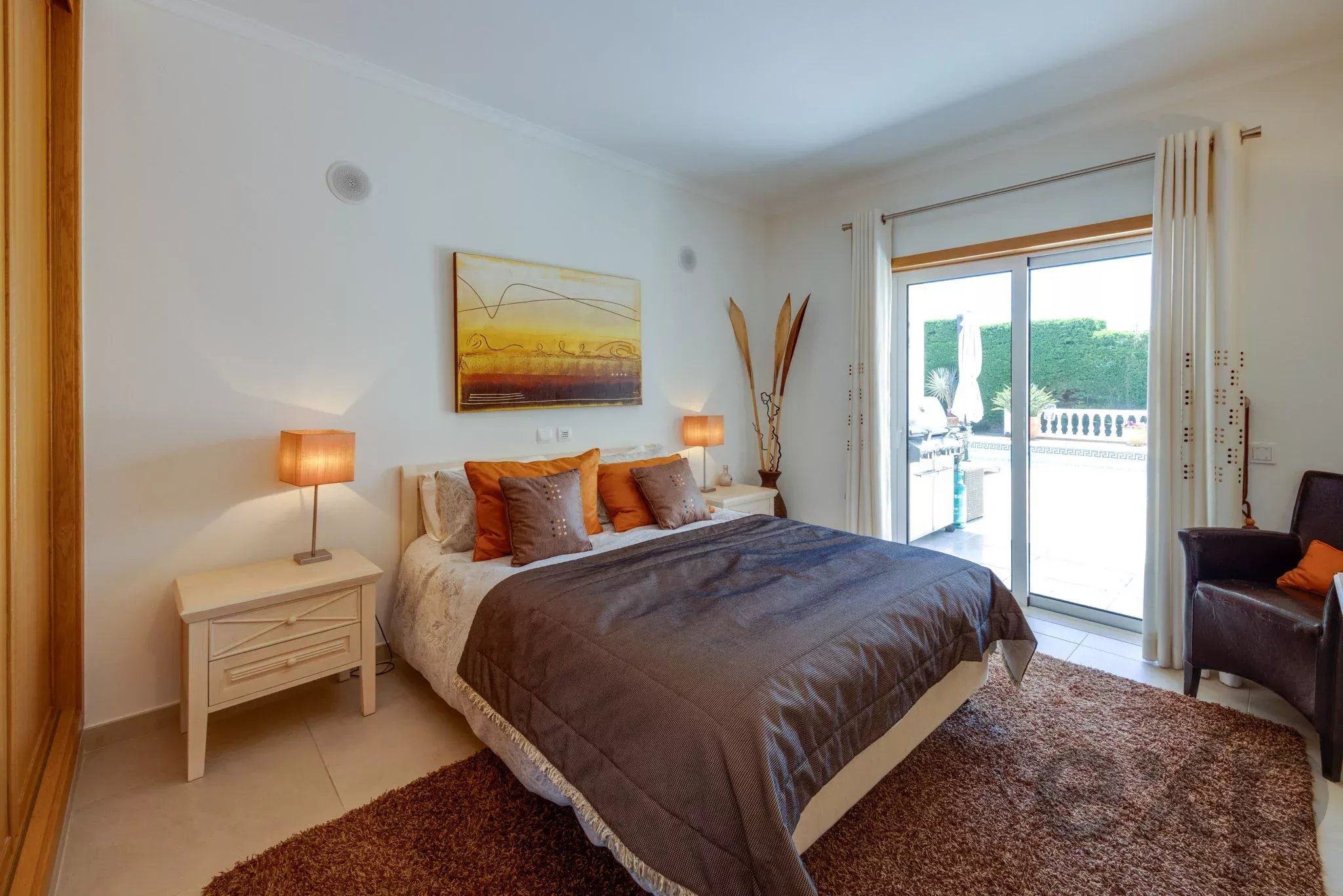 Bright hotel-style bedroom with a large bed, brown and gray pillows, and twin bedside lamps. A sliding glass door opens to a sunny balcony garden; white walls and abstract wall art.