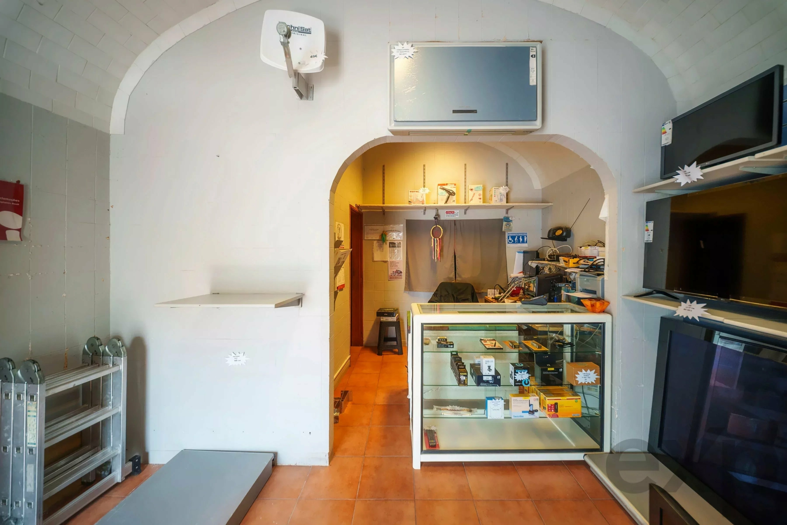 Interior of a small shop with a glass display counter filled with electronics and accessories, white walls, and ceiling arch