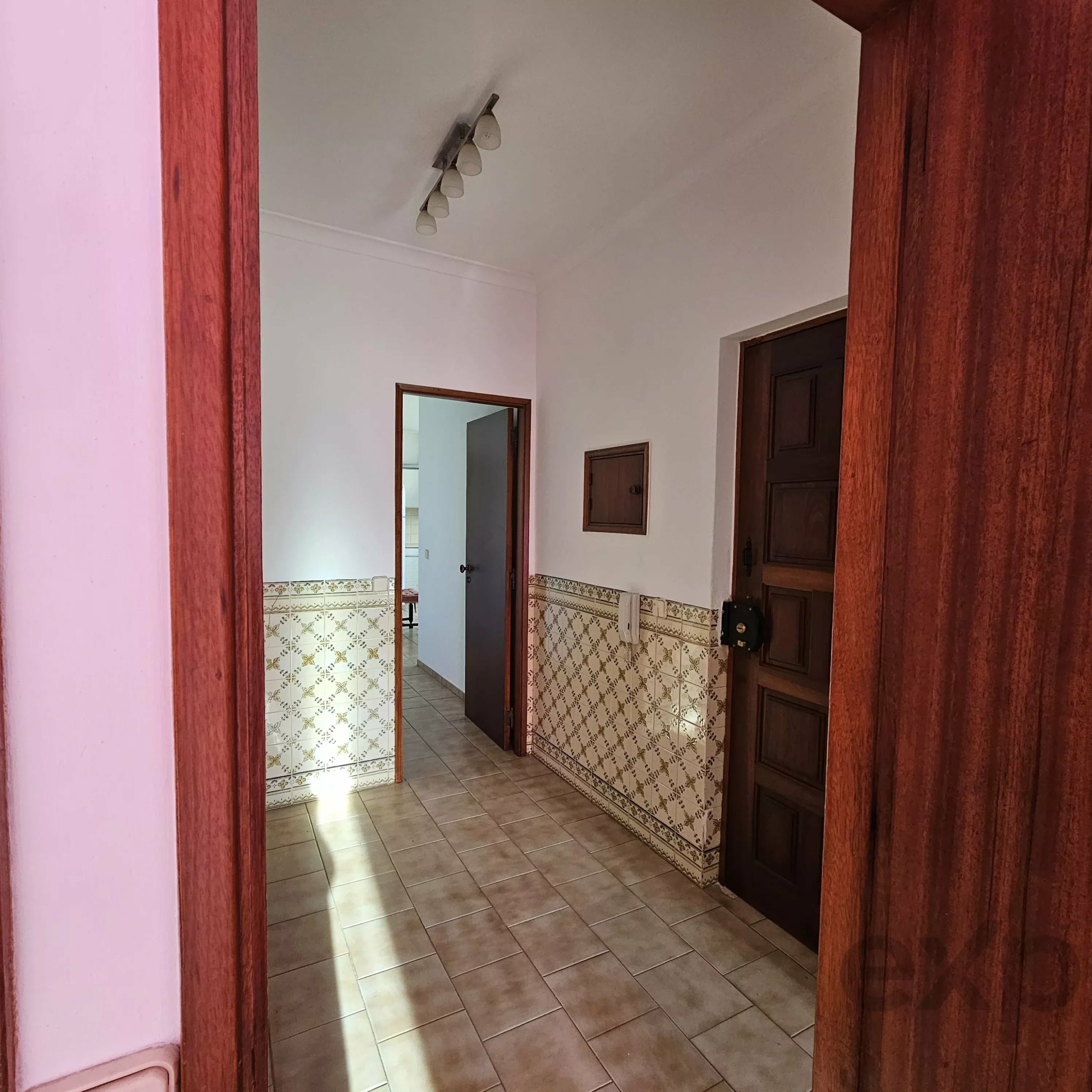 Interior doorway view into a tiled hallway with decorative tile wainscoting and a row of ceiling lights on a sloped ceiling