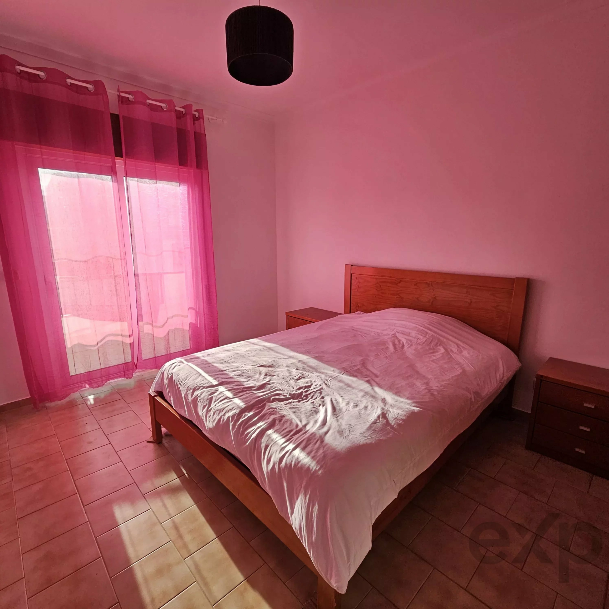 Twin wooden beds with white bedding in a pink-lit bedroom, pink sheer curtains by a window on the left
