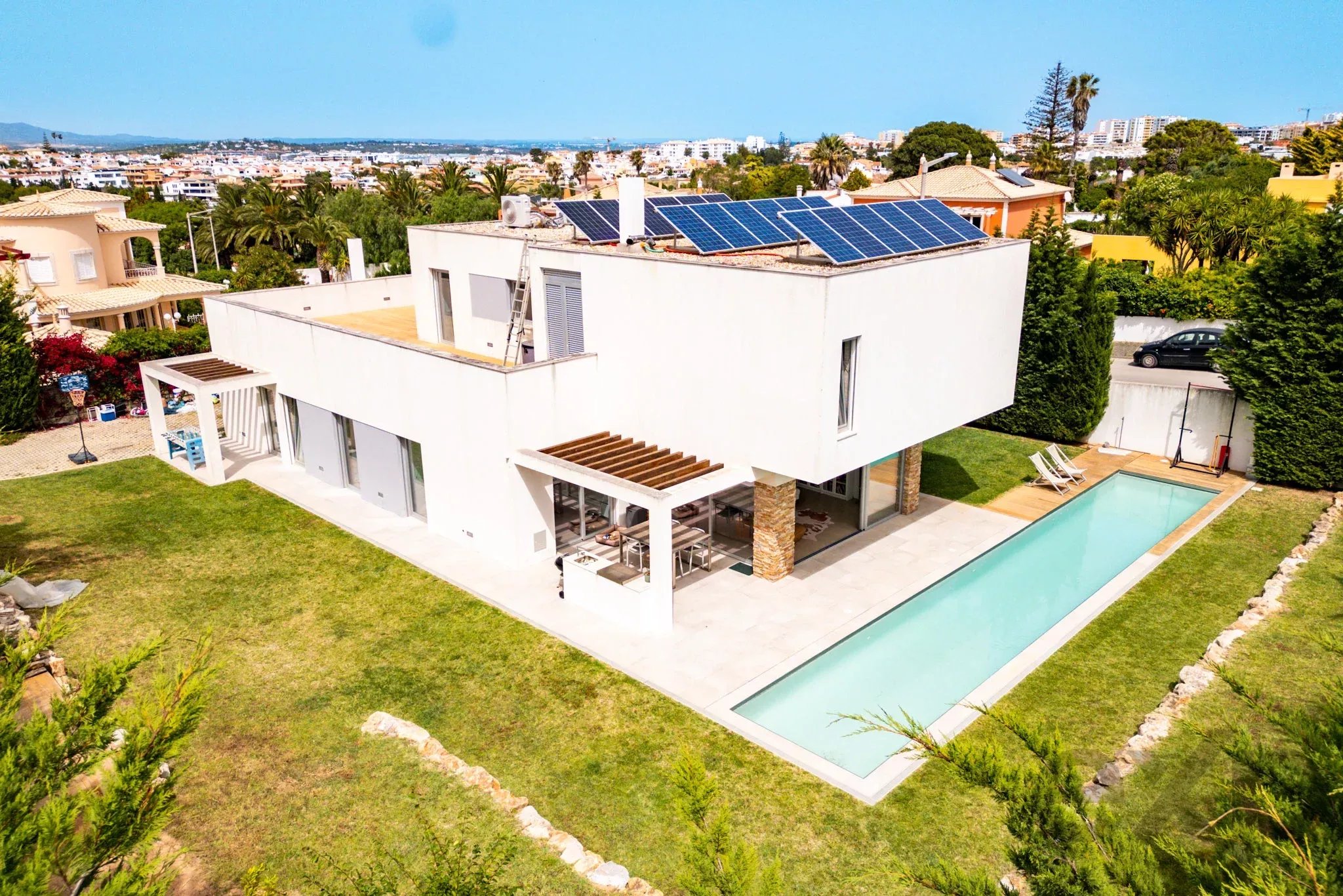 Aerial view of a modern white house with solar panels on the flat roof, outdoor seating area, and a long rectangular pool surrounded by green lawn.