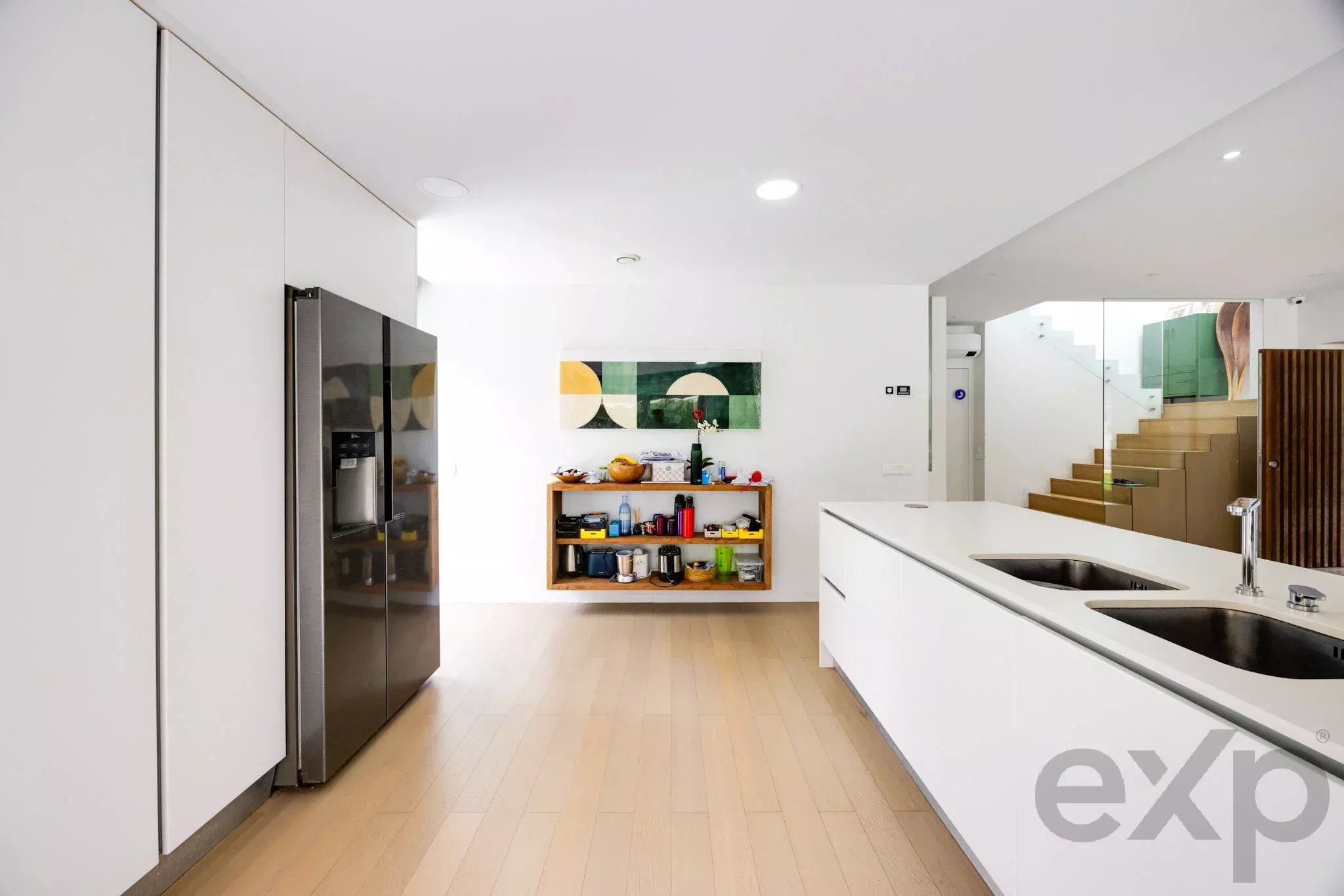 Modern white kitchen with a large island, built-in fridge, open shelving, and a staircase visible on the right.