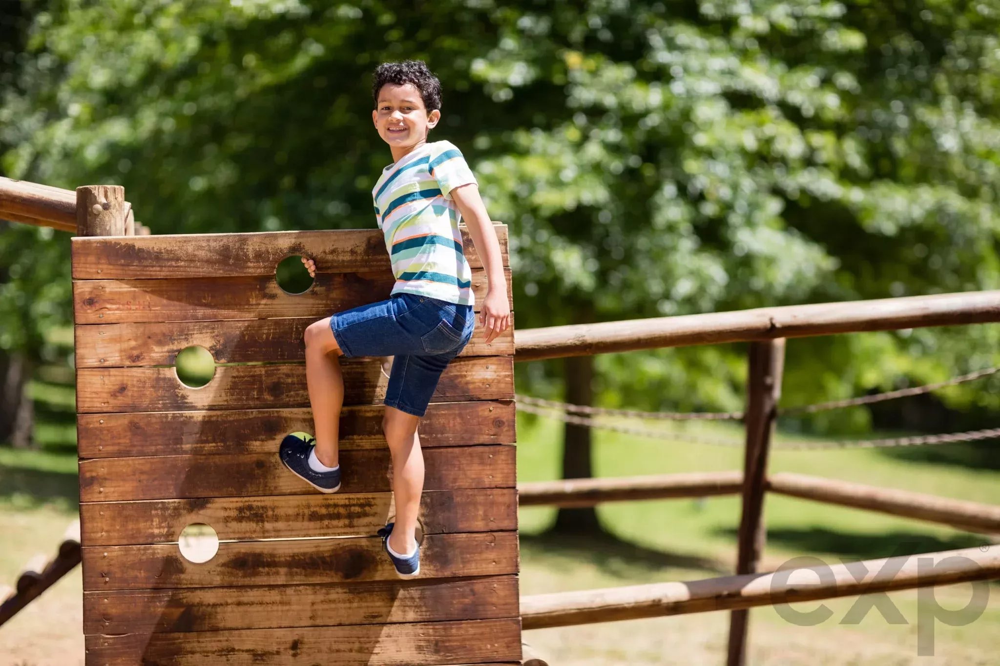Smiling boy in a striped shirt climbs a wooden obstacle in a sunny park.
