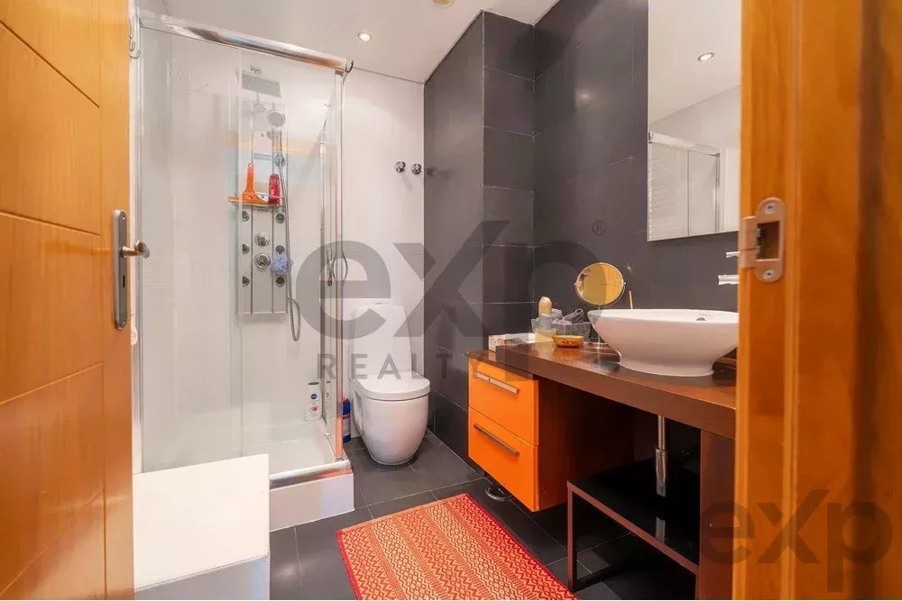 Modern bathroom with glass shower enclosure, toilet, and dark wall tiles; orange vanity drawers and white vessel sink on a wooden countertop.
