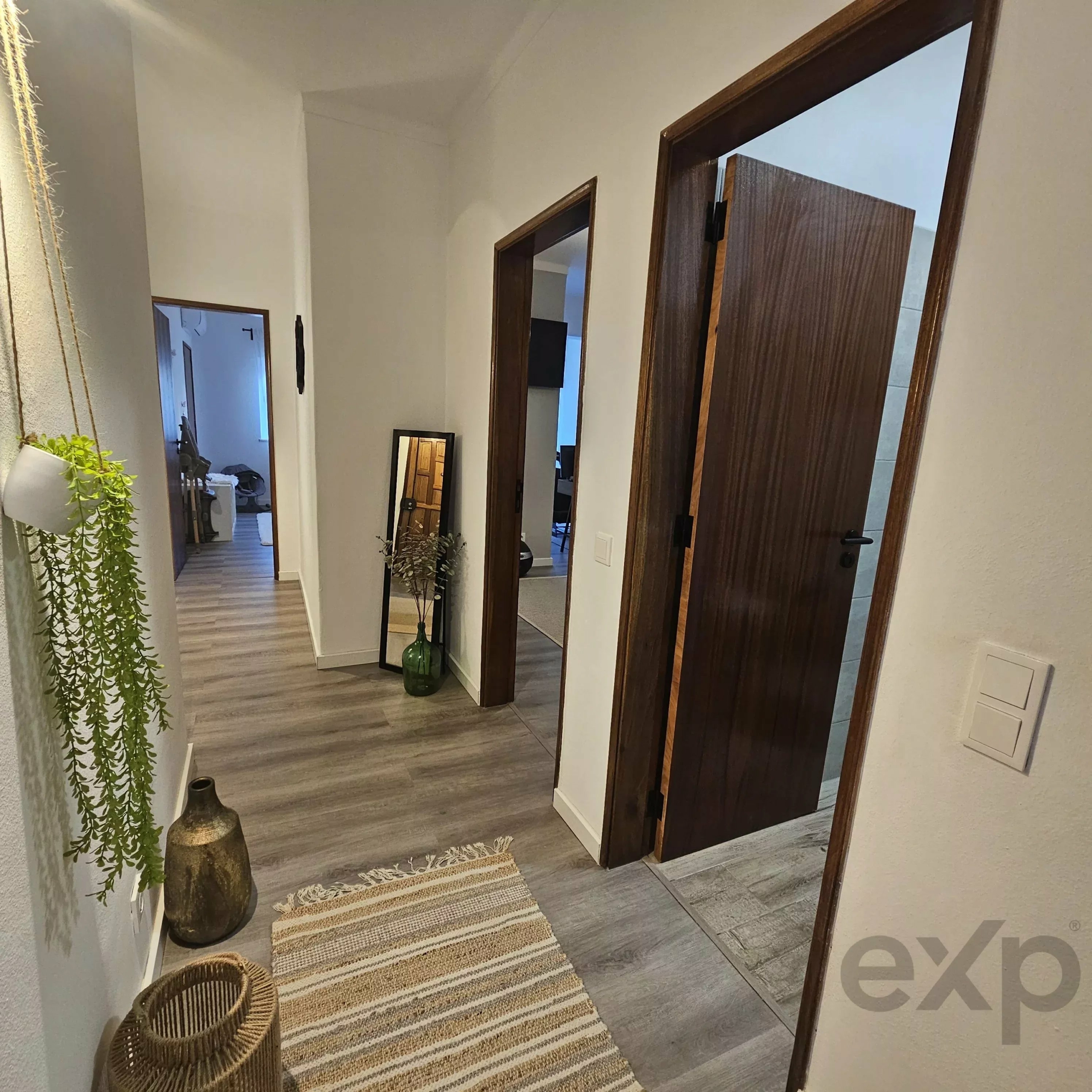 Narrow hallway with light walls, several open wooden doors, a striped woven rug, and a tall plant in a wicker basket at the end.