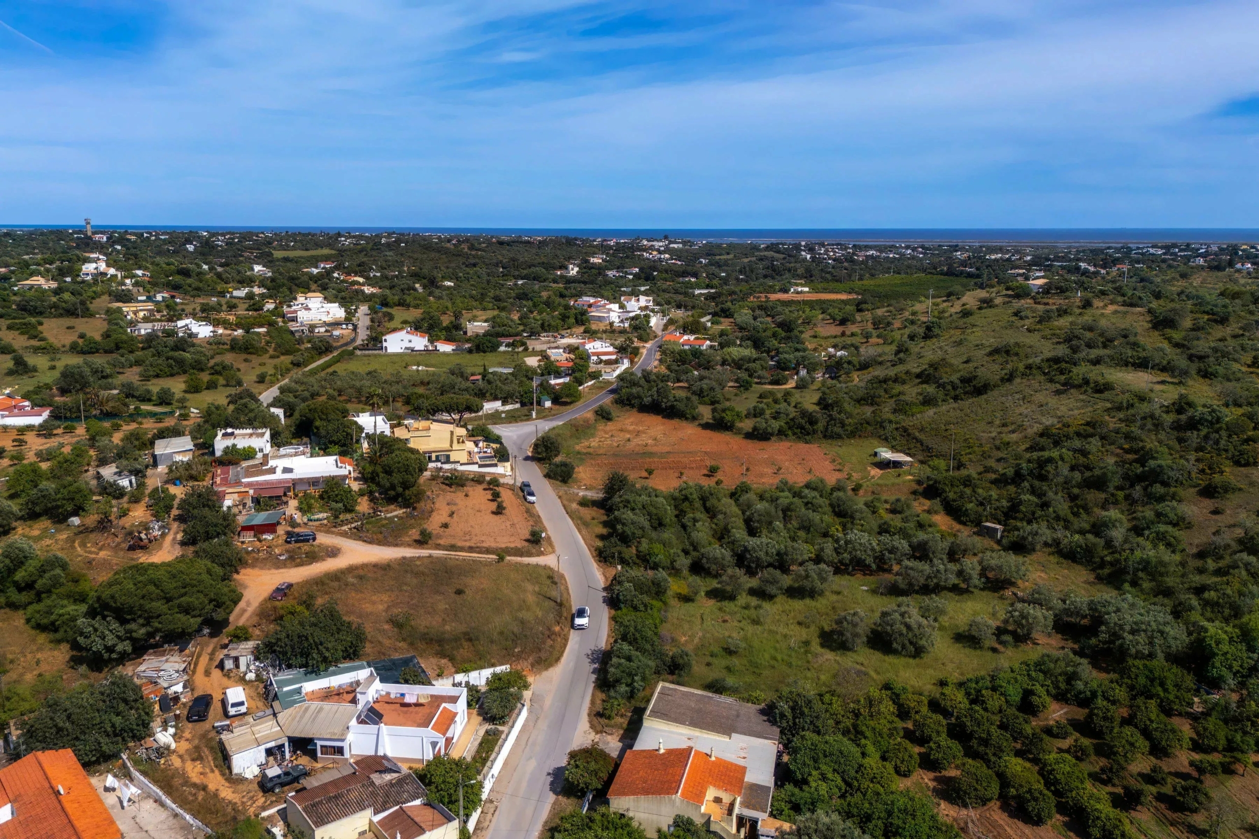 Aerial view of a small coastal town with white buildings, red roofs, and winding roads surrounded by trees and shrubland, ocean on the horizon.