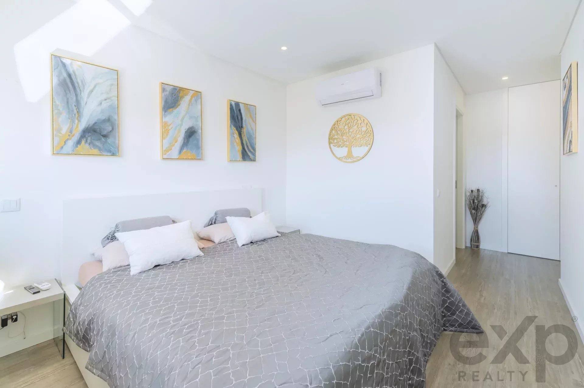 Bright bedroom with a large bed, gray patterned comforter, white pillows, and three abstract paintings with gold frames on the wall.