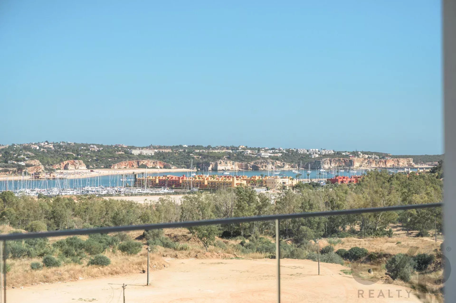 Harbor with numerous moored sailboats, colorful low-rise buildings along the shore, and hills in the distance under a clear blue sky, seen from a balcony railing.