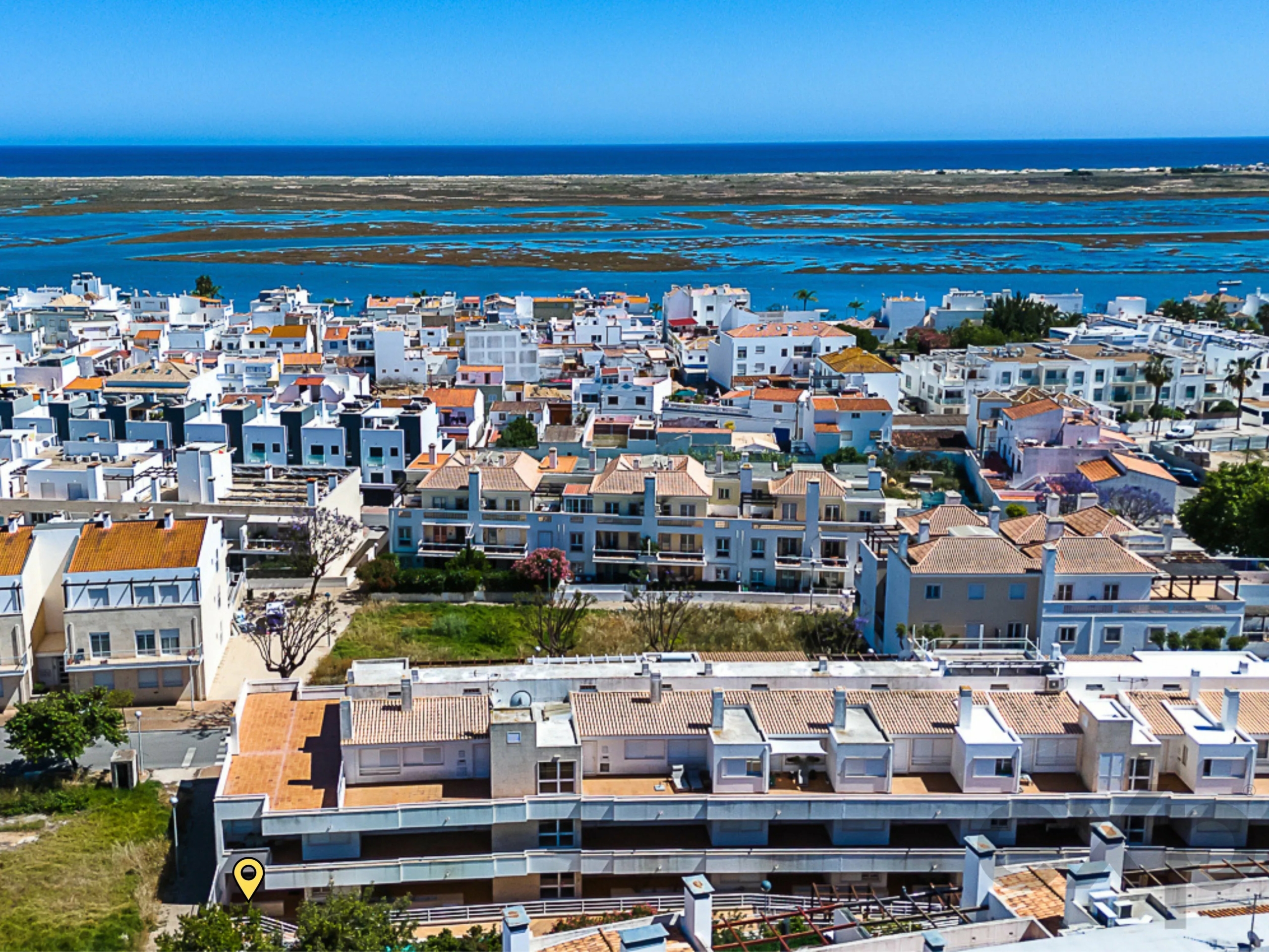 Aerial view of a white, Mediterranean-style coastal town with orange-tiled roofs, set against the blue sea and marshy coastline.
