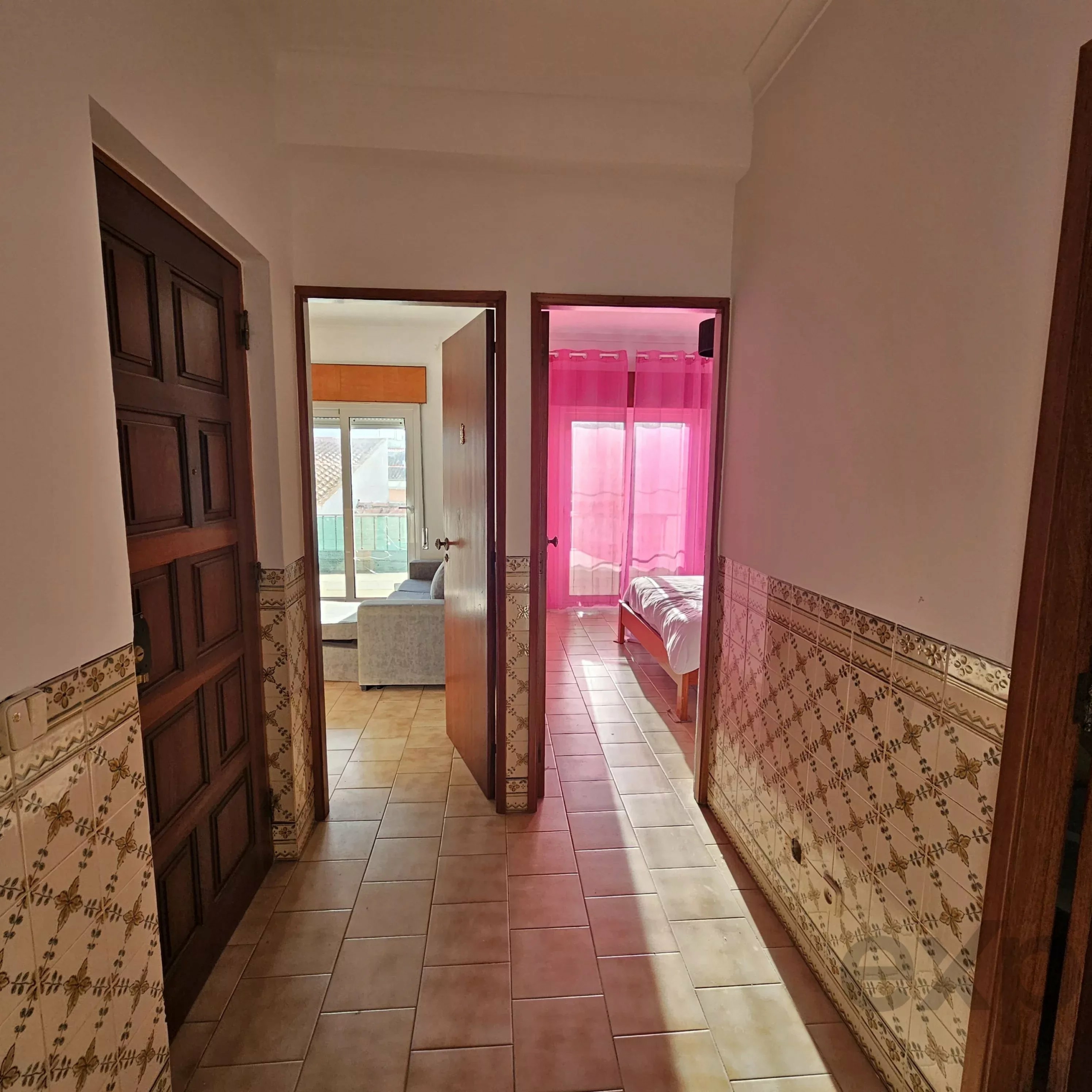 Narrow hallway with tiled floor and wooden doors, one open to reveal a pink-curtained room beyond.