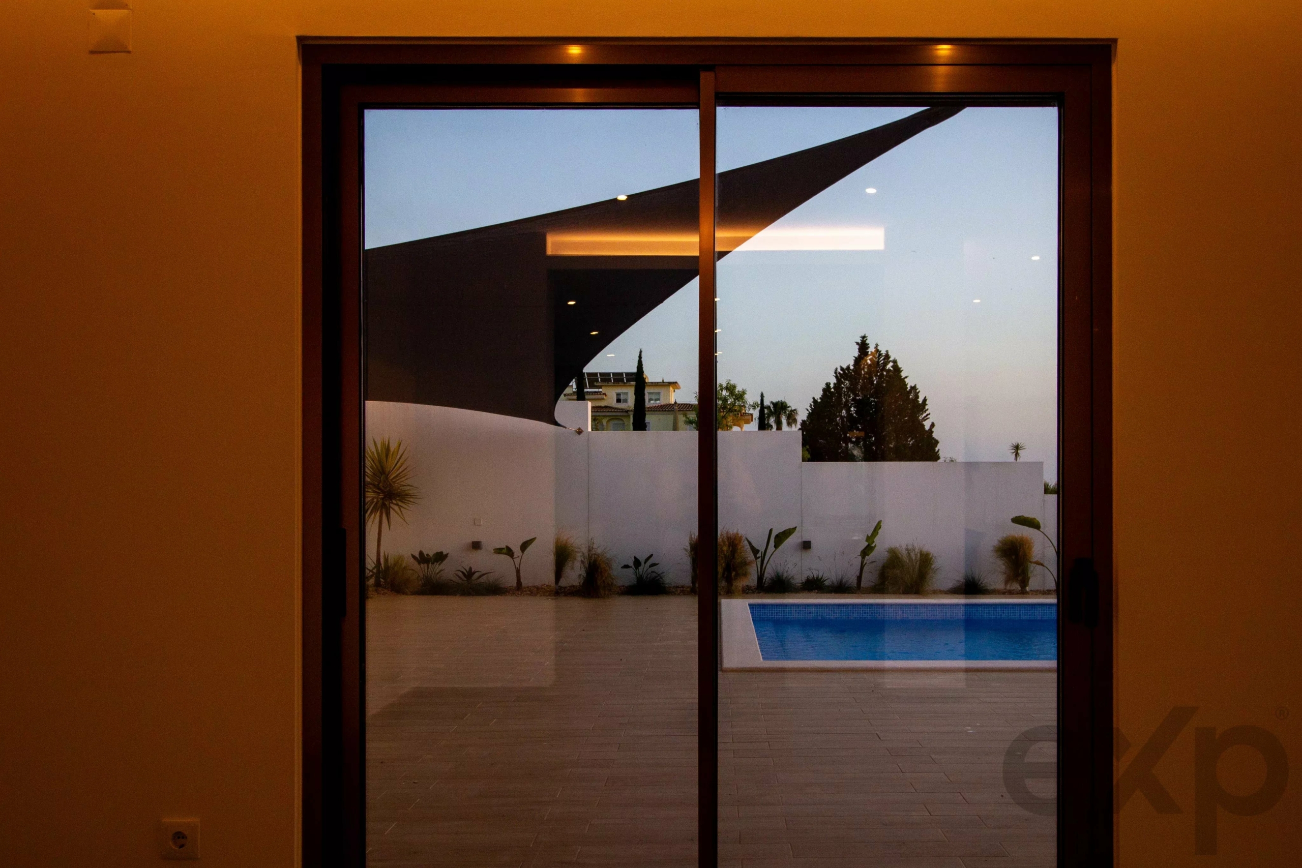 View through glass doors to a backyard with a swimming pool, white privacy wall, and a large diagonal shade sail against a blue evening sky.