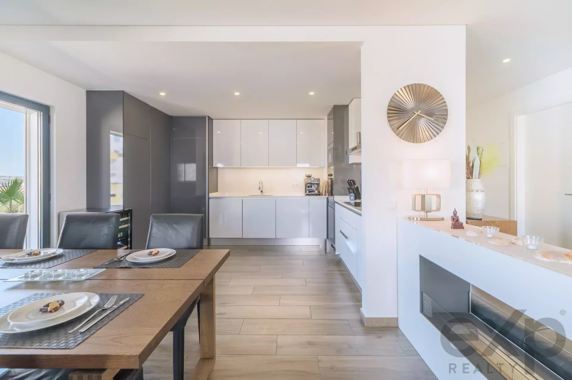 Open-plan kitchen and dining area with white and gray cabinetry, a wooden dining table set for four, and a decorative wall clock nearby.