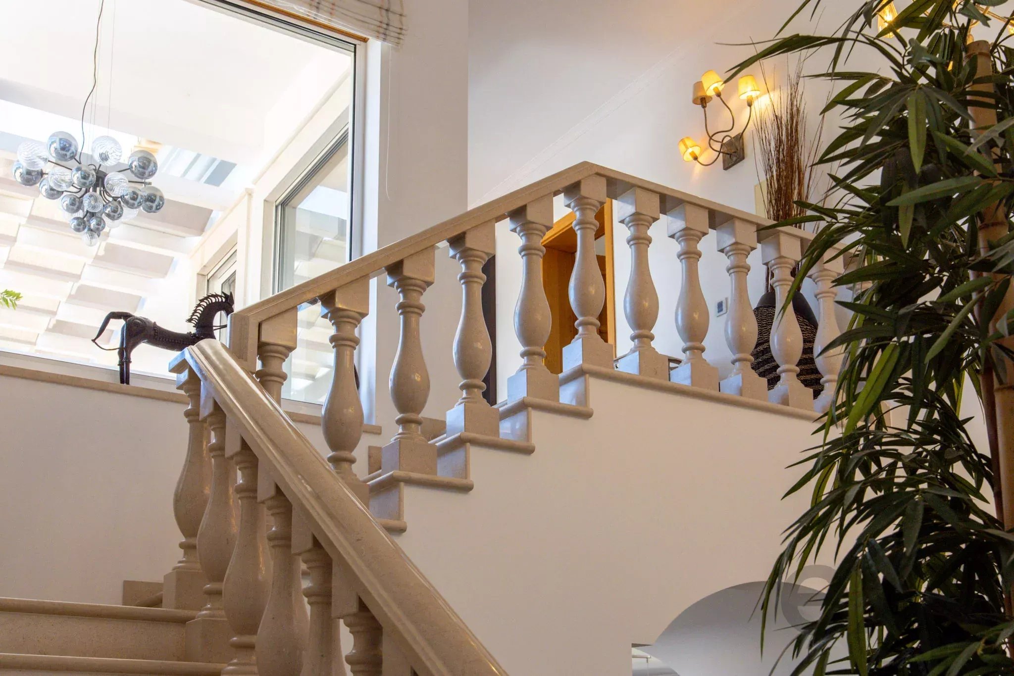 Indoor staircase with a beige balustrade and handrail, glass-sphere chandelier on the left, and tall green plants on the right.