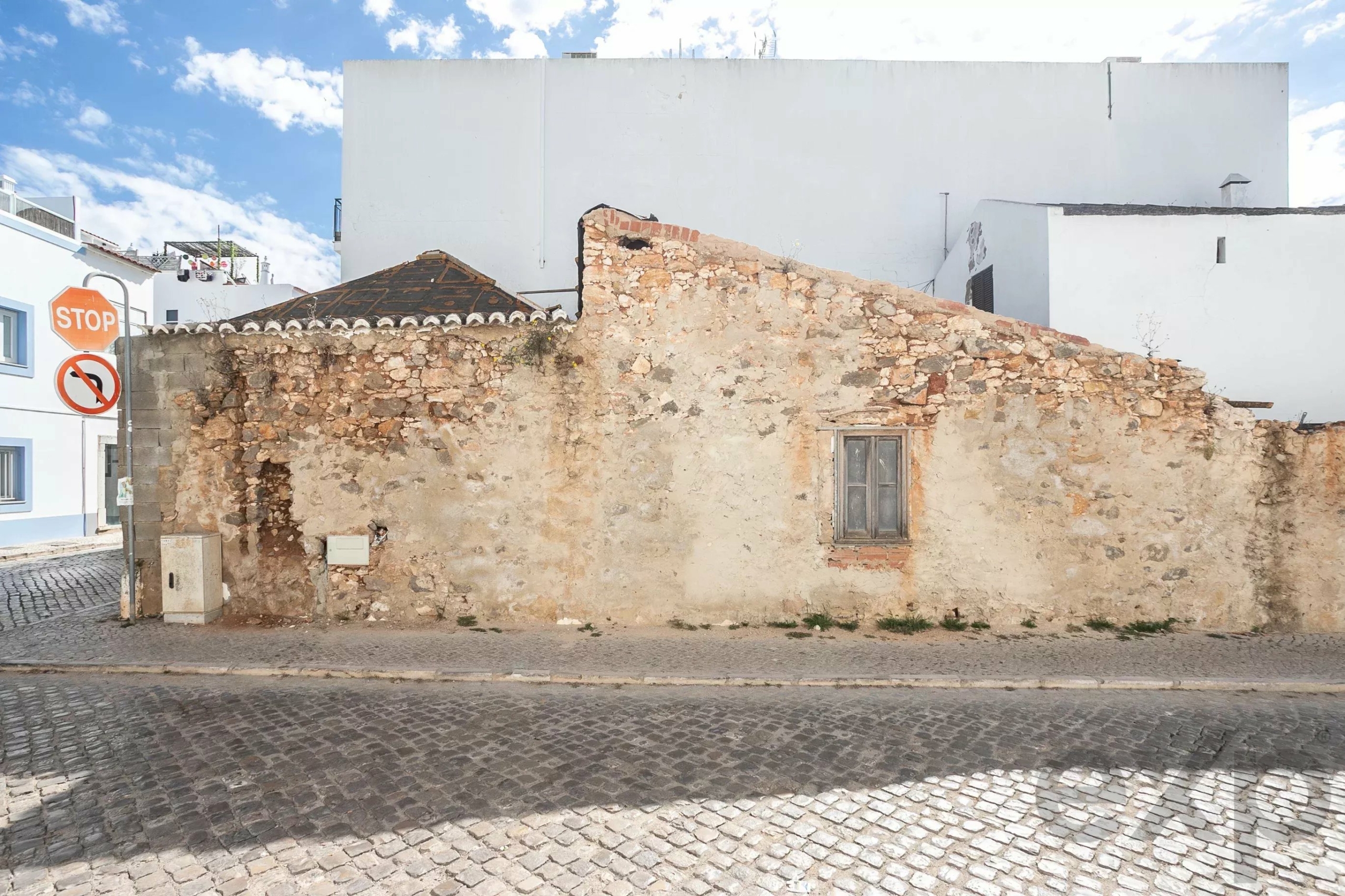 Old stone wall along a cobblestone street with a Stop sign and white buildings in the background under a blue sky.