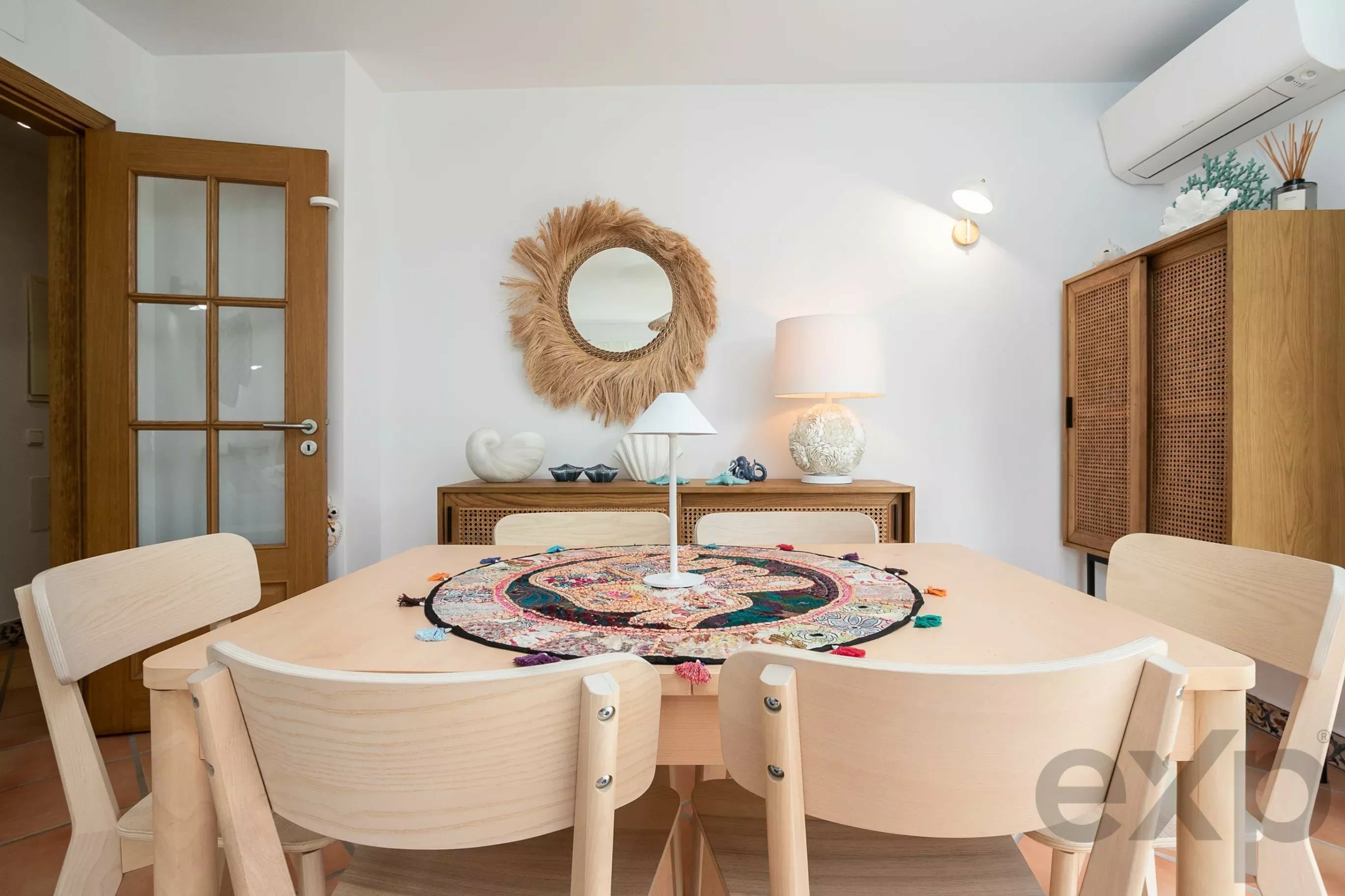 Dining room with light wood table and chairs, a colorful round tassel table runner, and a woven-framed mirror on a white wall.