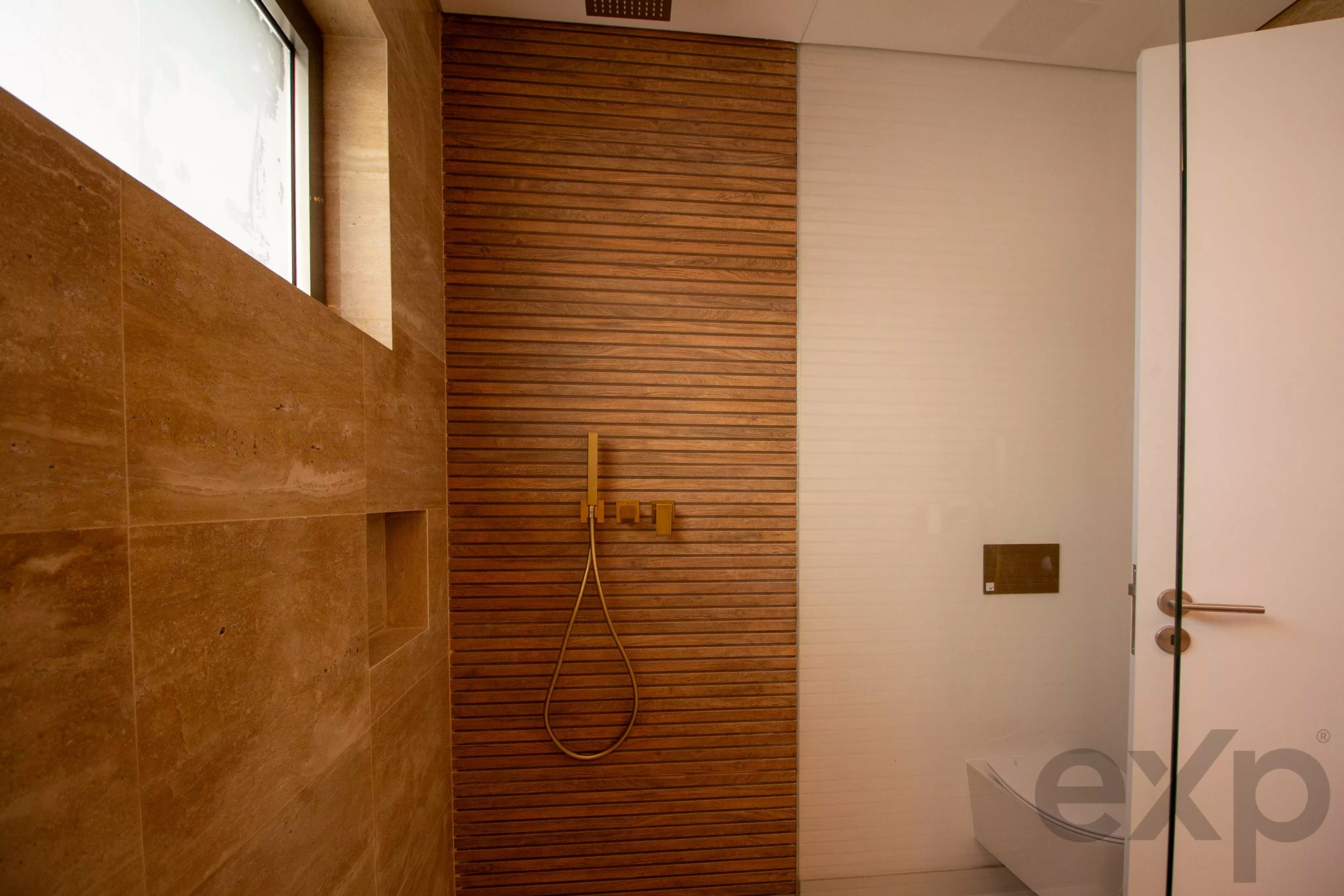 Shower area with a vertical wood-slatted wall, beige stone tiles, and a brass handheld shower on a hose. A glass door is visible on the right.