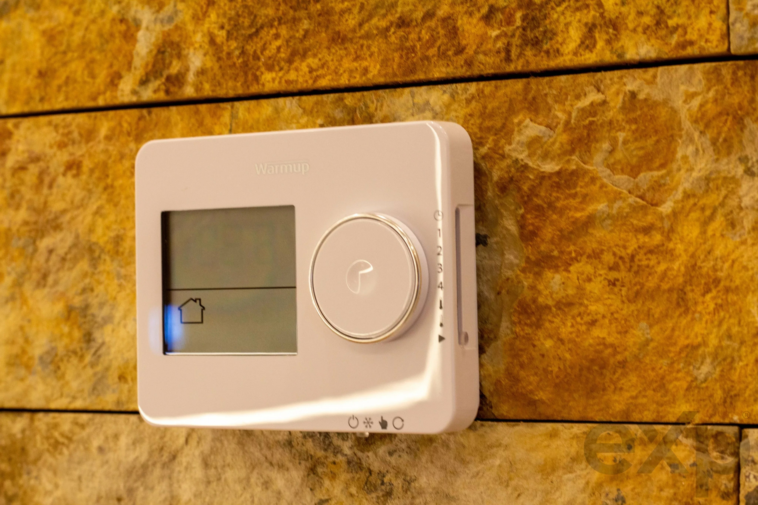 White wall-mounted thermostat with LCD screen and large circular dial on a textured brown stone wall