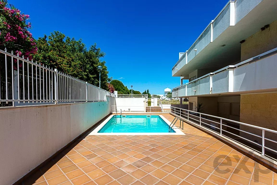 Rectangular outdoor swimming pool surrounded by terracotta tiles, white railings, and a modern apartment building under a clear blue sky.