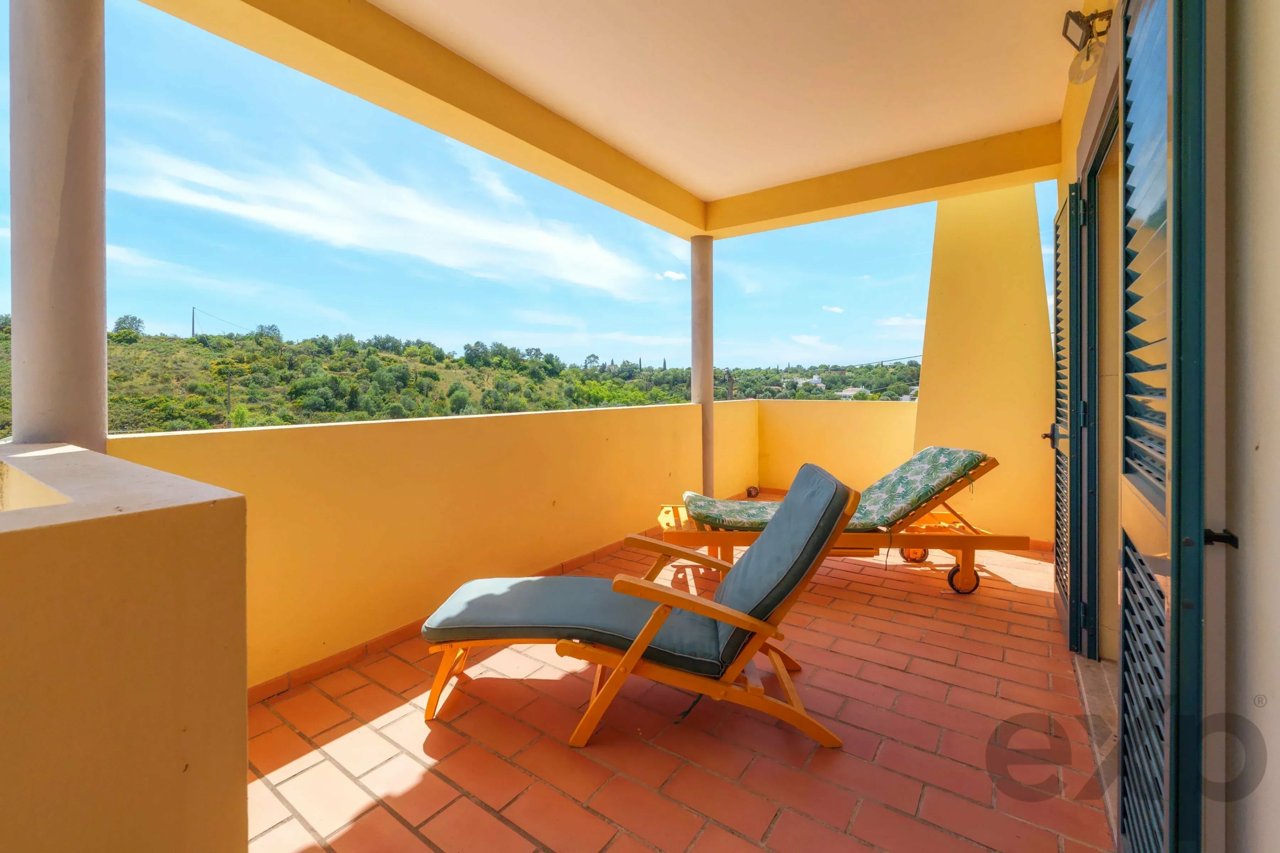 Sunlit yellow balcony with terracotta tiles and two green-cushioned lounge chairs facing a hillside landscape under a blue sky