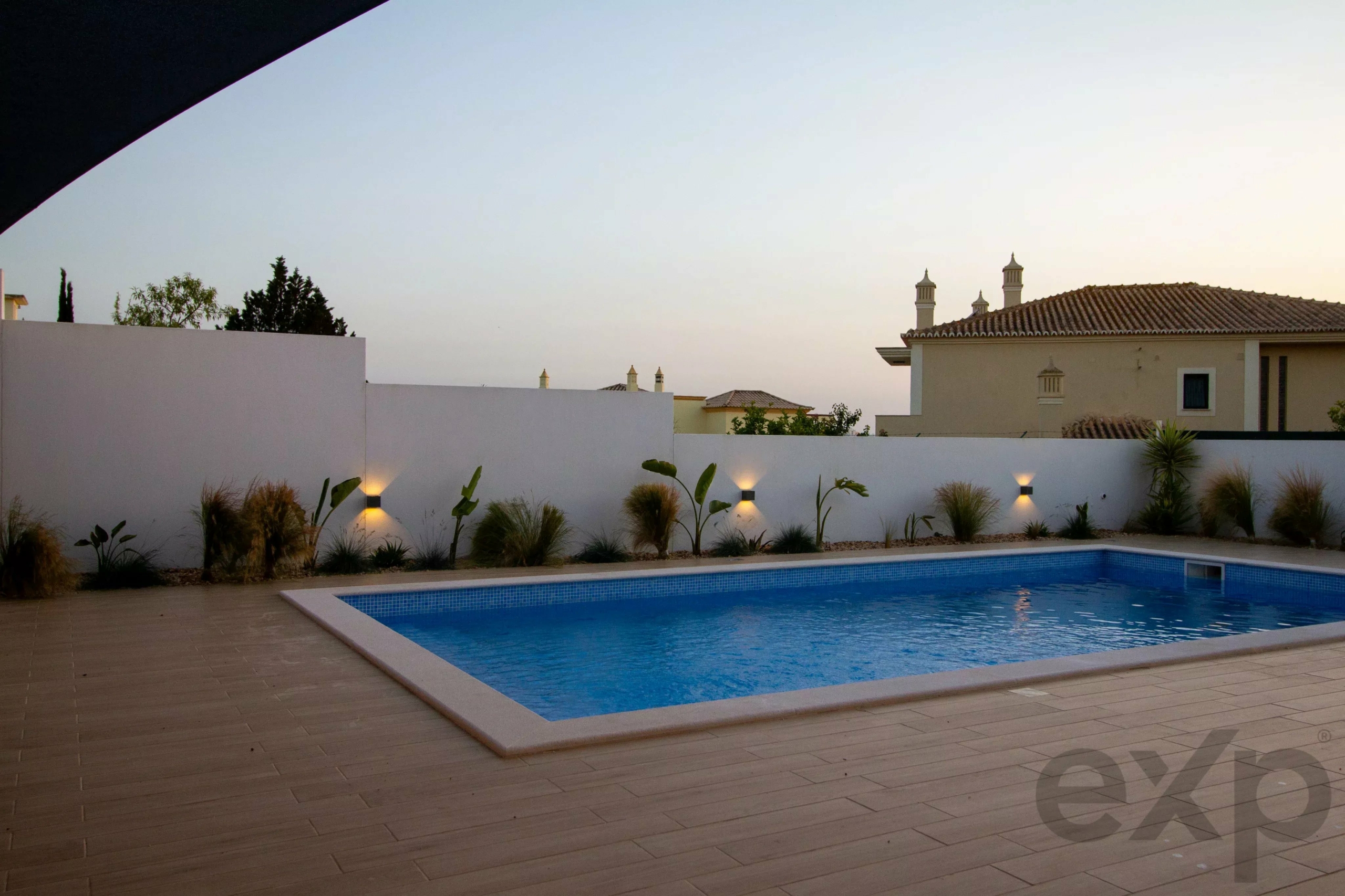 Rectangular blue swimming pool in a modern courtyard, with a beige tile deck and a white privacy wall; villas with tiled roofs in the distance at dusk.