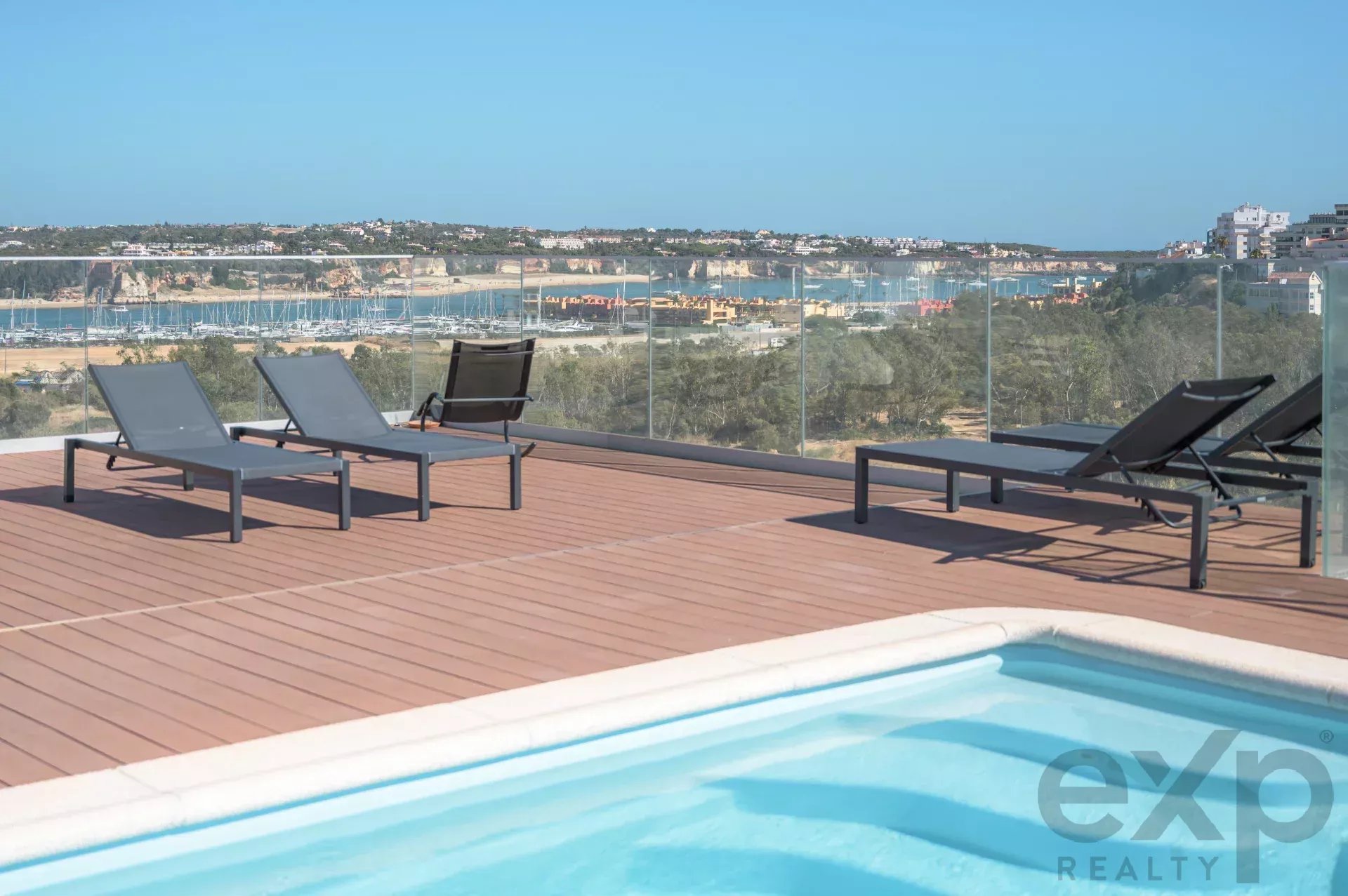 Poolside deck with grey lounge chairs on wooden planks, glass railing, overlooking a marina and coastal town.