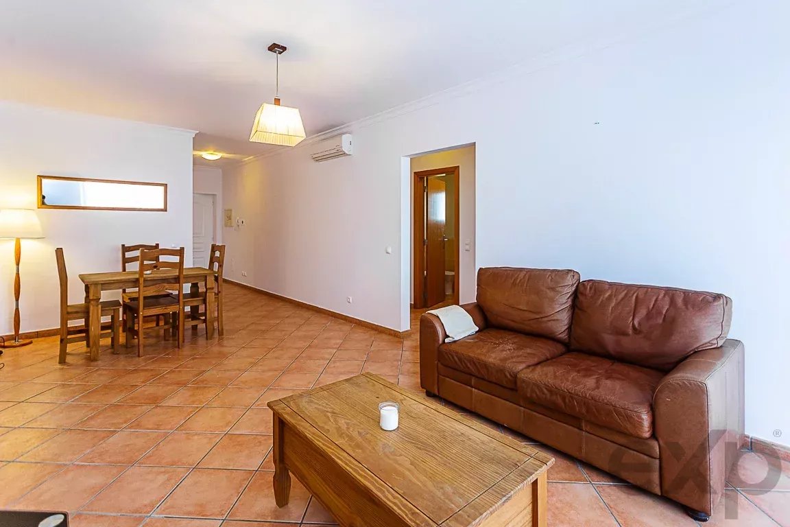 Living room with a brown leather sofa, wooden coffee table, and a dining table with chairs on a terracotta tile floor.