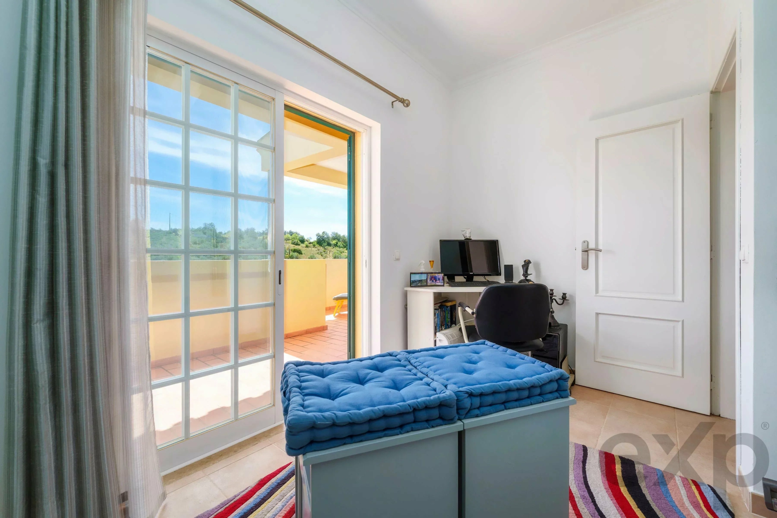 Bedroom with a bright, sliding glass door opening to a sunny balcony; a blue tufted bench sits at the foot of the bed and a desk with computer is against the opposite wall.
