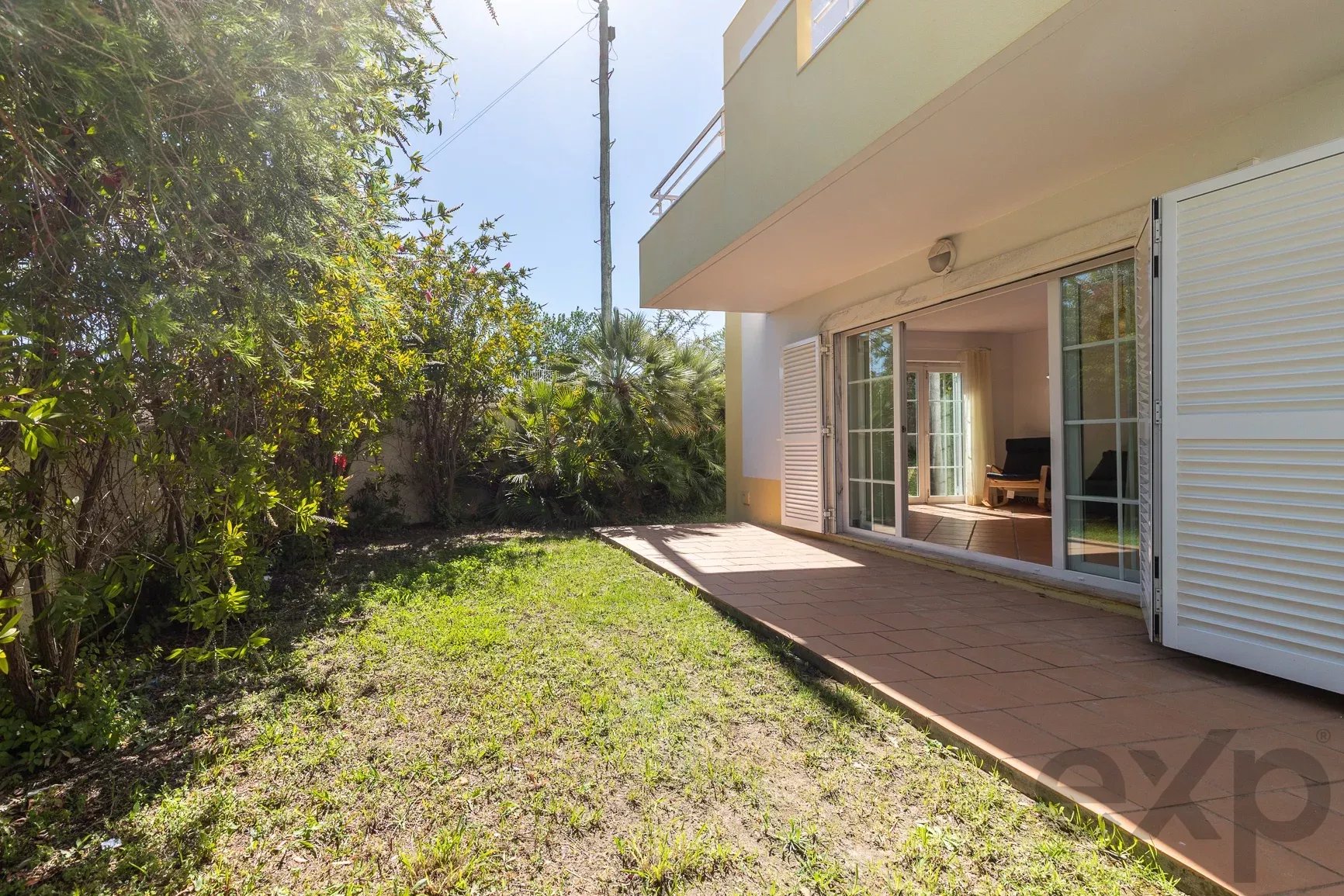 Backyard of a modern white house with an open sliding glass door leading to a tiled patio and green shrubs nearby.