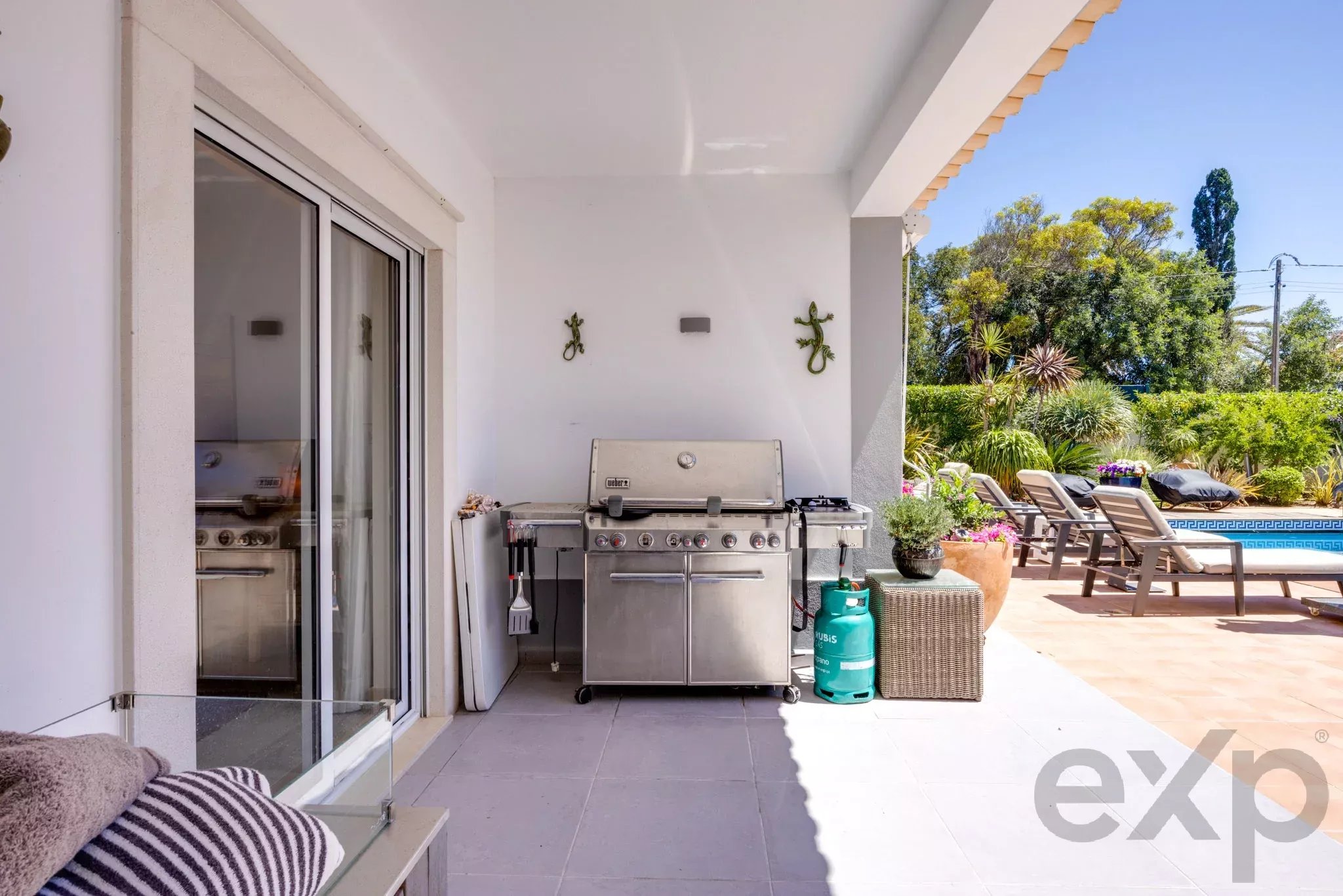 Outdoor patio with built-in barbecue grill, sliding glass door, and lounge chairs beside a pool and garden.