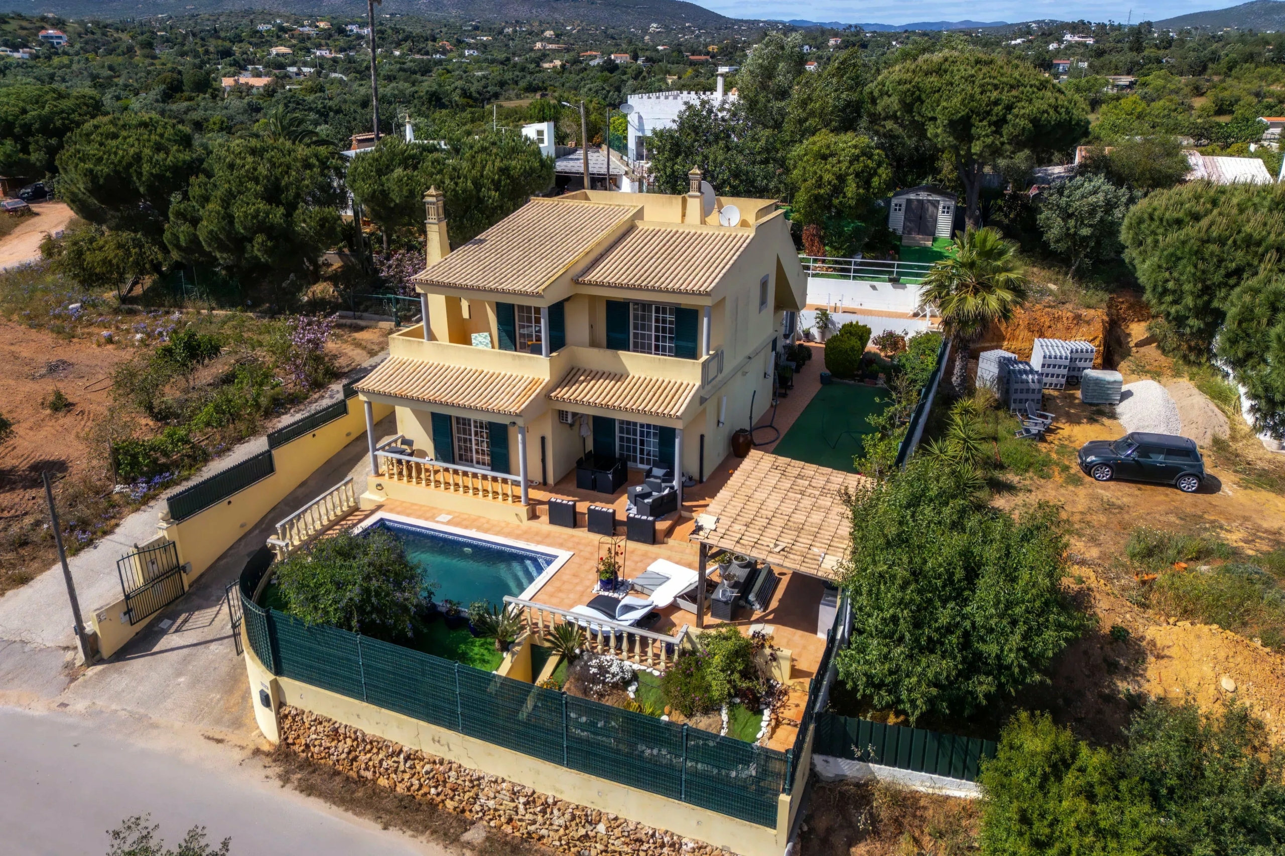 A two-story yellow villa with a tiled roof and a wraparound terrace, pool in the foreground, surrounded by a green garden and hillside neighborhood.